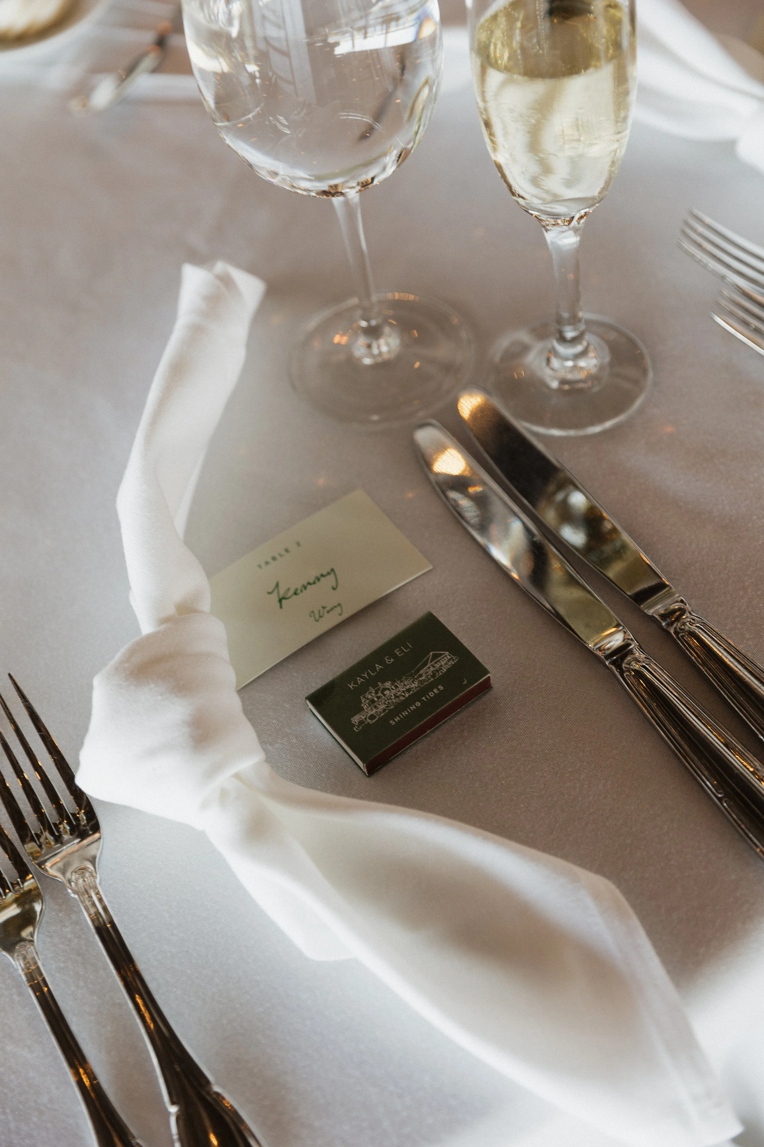 A formal table setting with two wine glasses, a white cloth napkin wrapped around a silverware, a green place card, a matchbox, and silverware arranged on a white tablecloth.