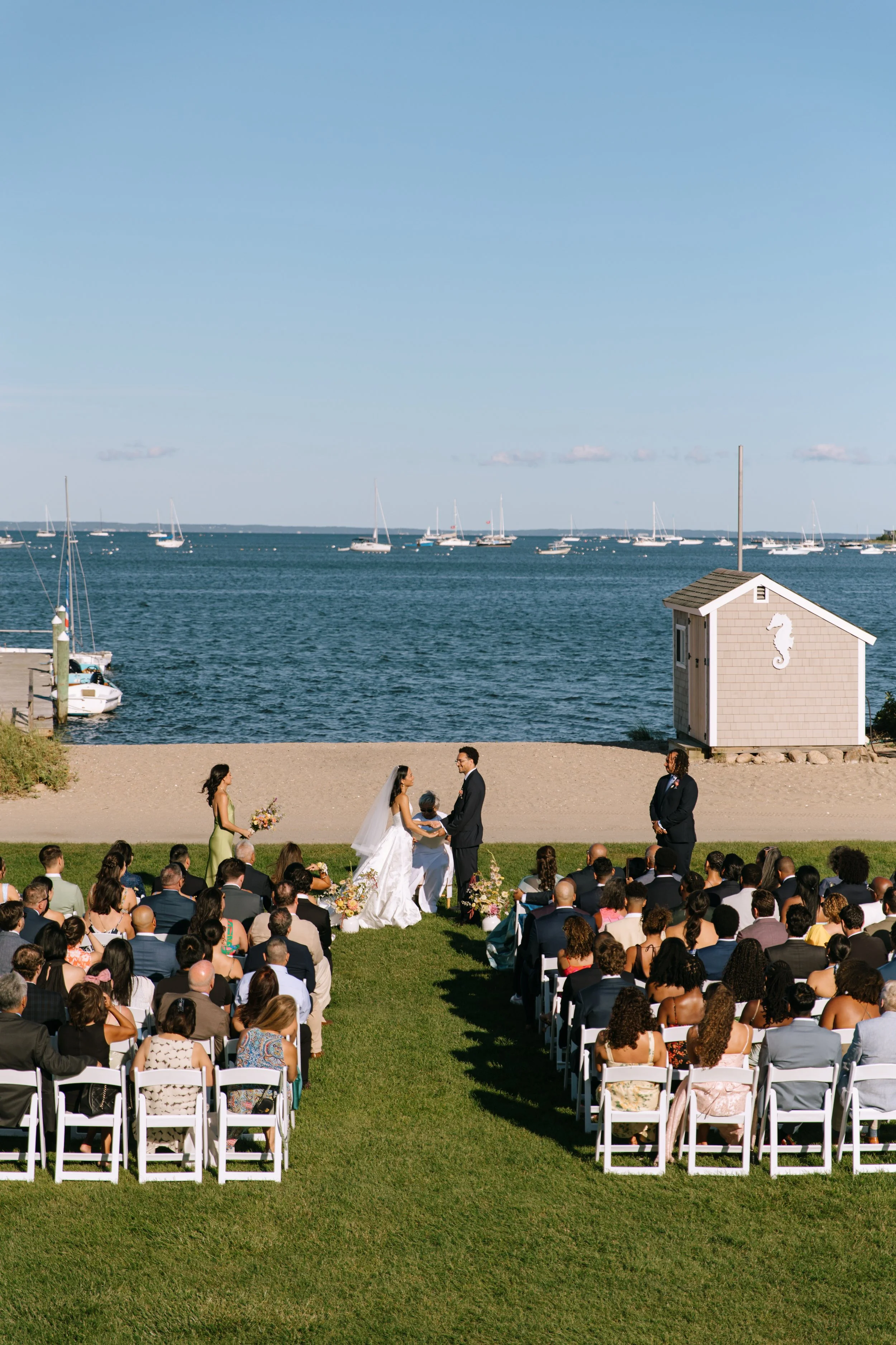 A wedding ceremony taking place outdoors on a lawn by the water with boats anchored in the background.
