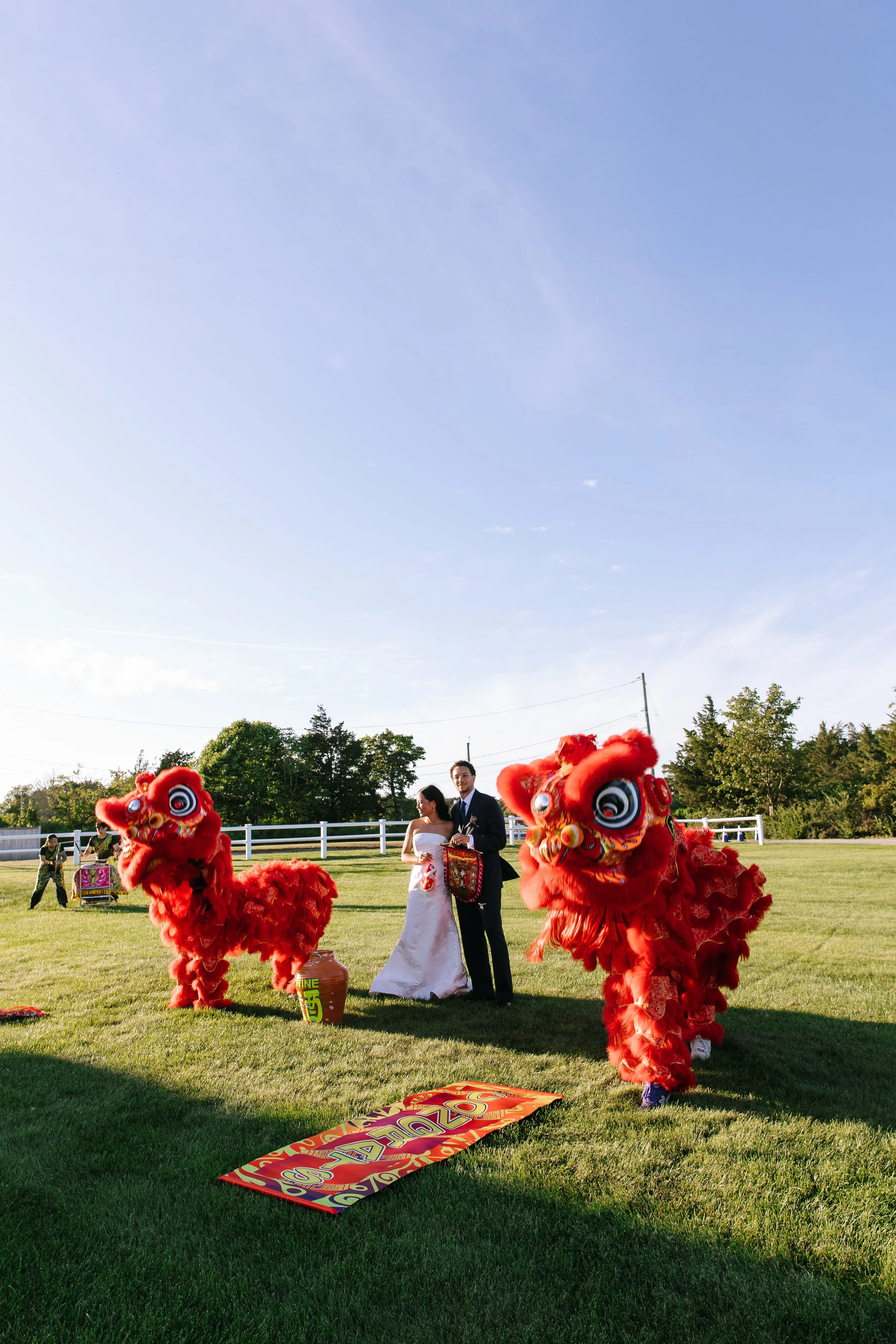 A bride and groom at a wedding celebrating with two red lion dance performers outdoors on a grassy field during daytime.