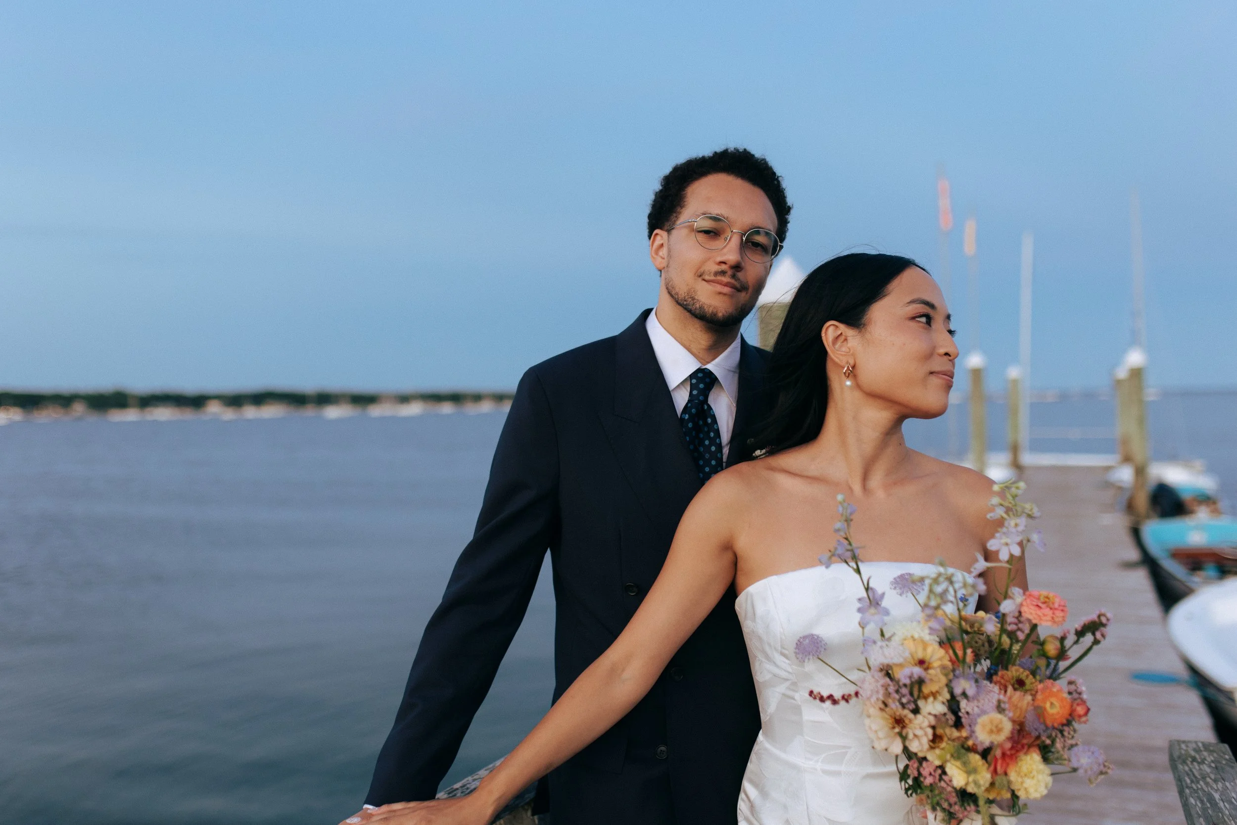 A couple dressed in wedding attire standing on a pier by the water, with a bouquet of flowers and boats in the background.