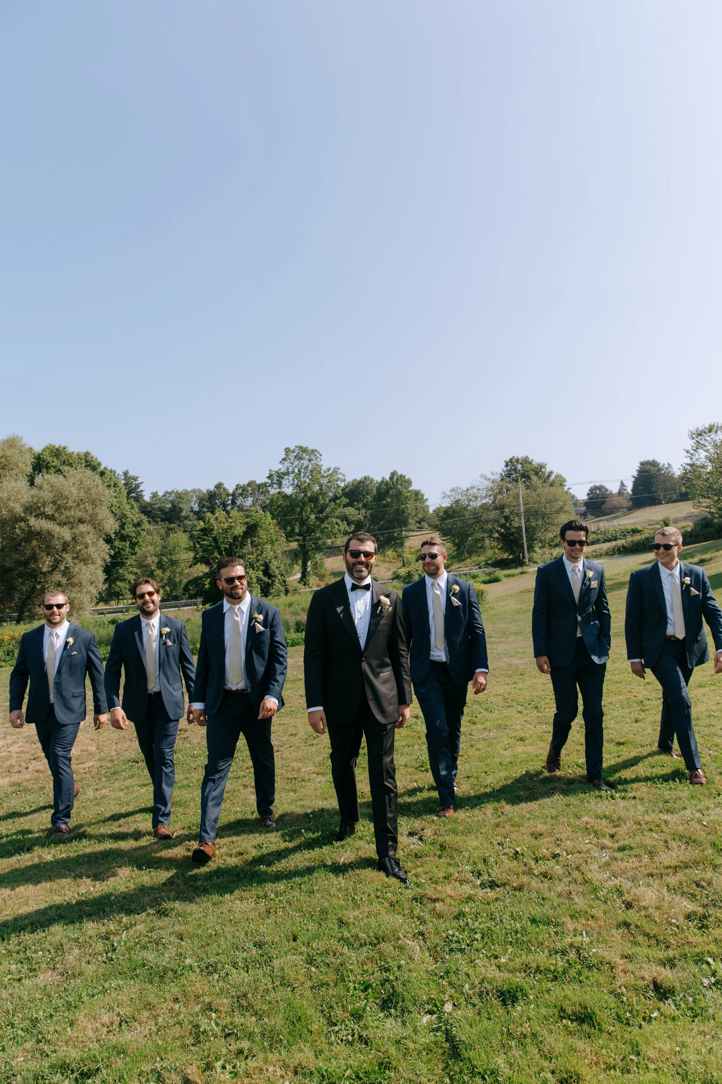 Groom and groomsmen walking on grass lawn during daytime outdoor wedding with trees and blue sky.