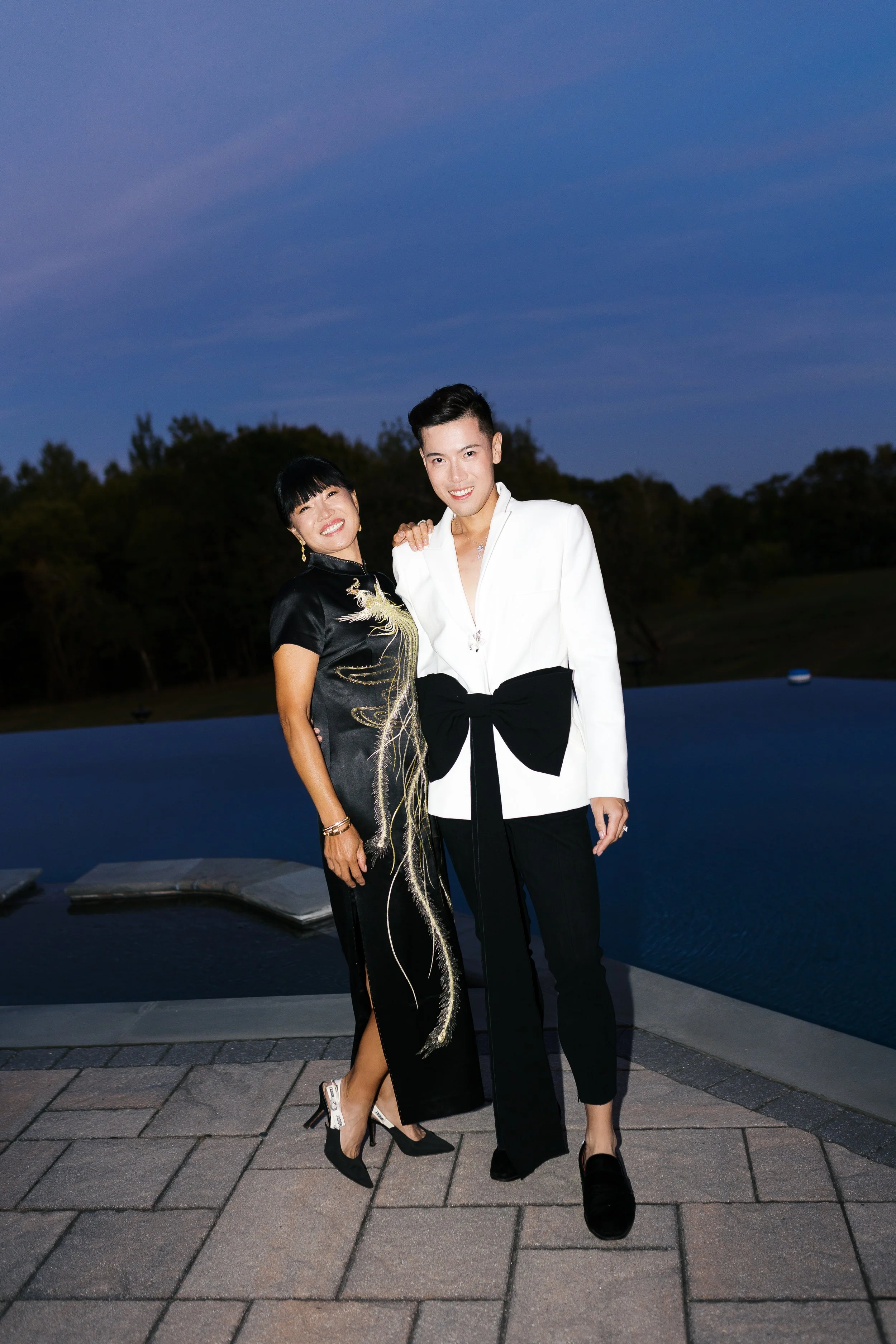 Two people, a man and a woman, standing together outdoors near a swimming pool during evening time, smiling at the camera. The woman is wearing a black dress with gold embroidery, and the man is dressed in a white blazer with a black bow detail and b