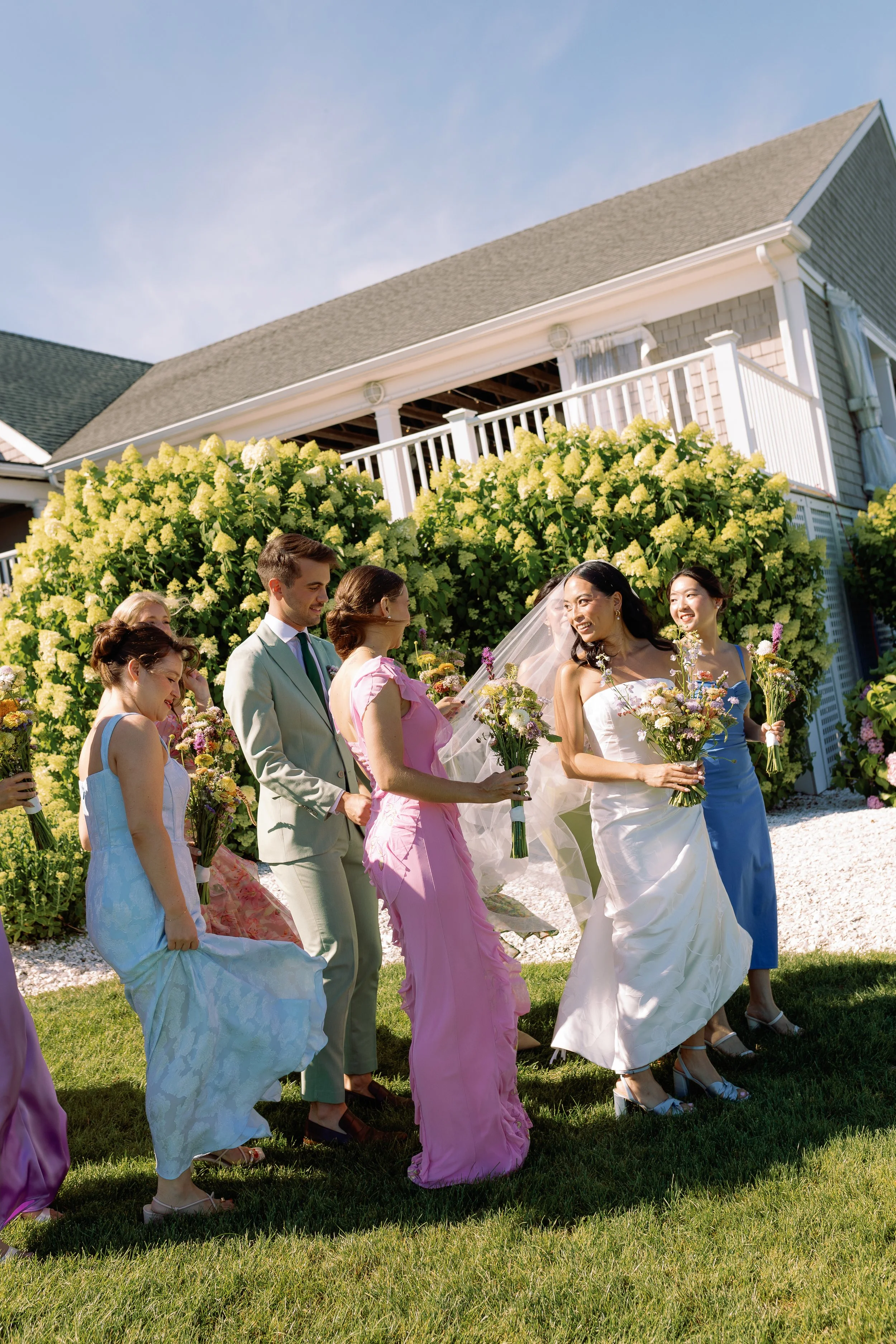 A bridal party gathering outdoors near a house with yellow hydrangea bushes, with women in colorful dresses and a man in a suit, holding bouquets of flowers.