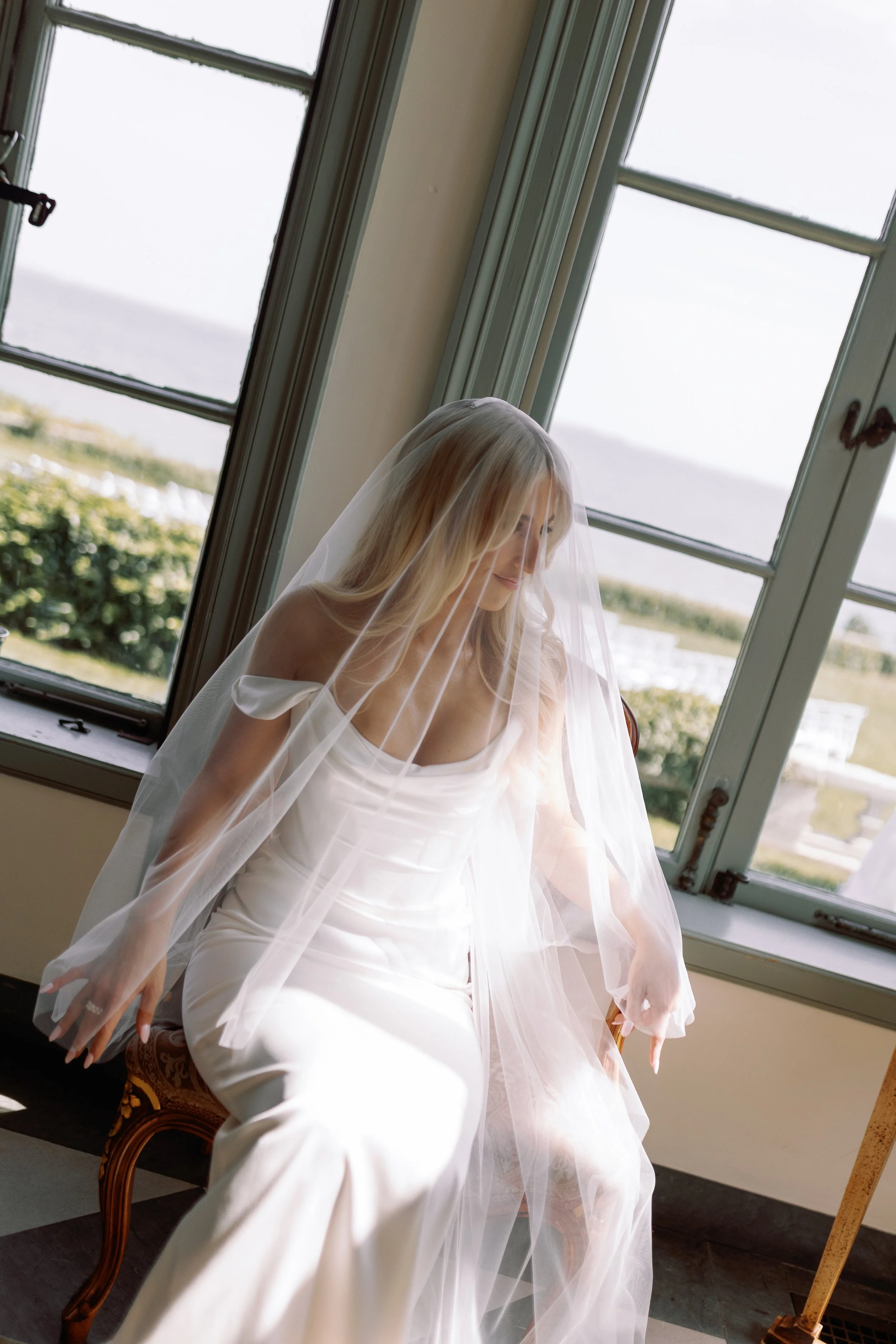 A woman with long blonde hair in a white wedding dress and veil sitting on a vintage wooden chair near large window, looking down, with sunlight shining on her.