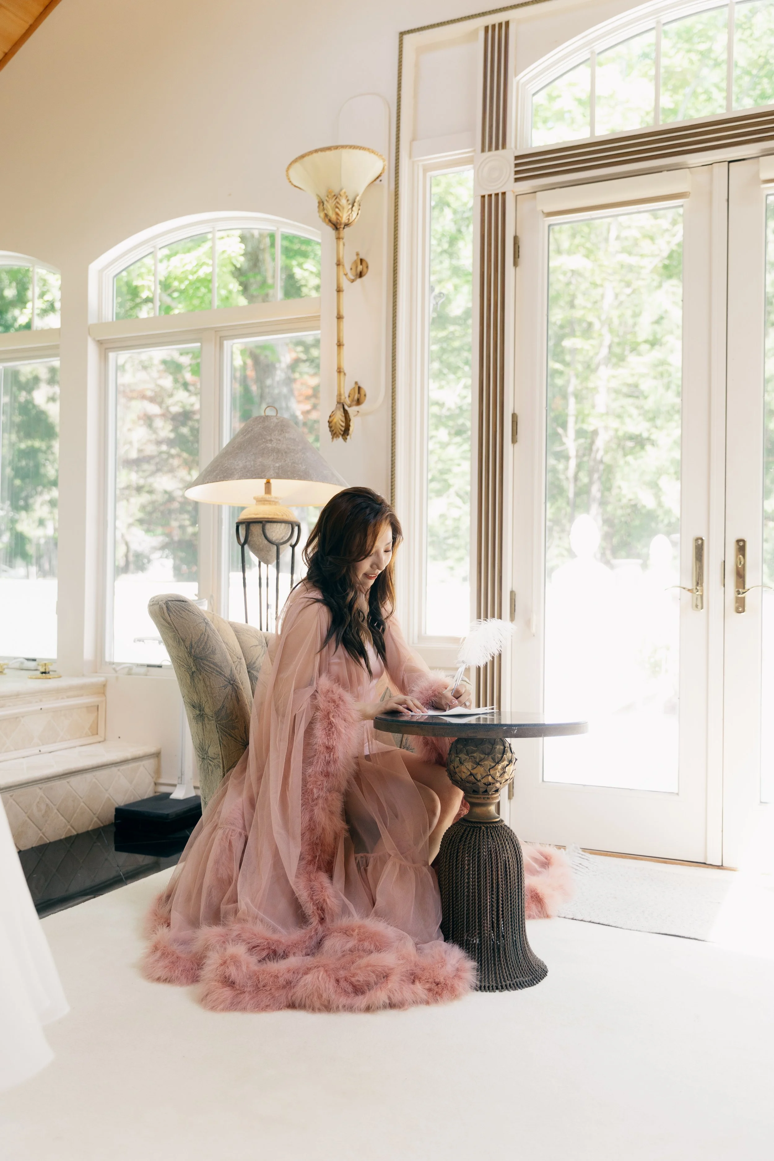 A woman in a pink, feather-trimmed gown sits at a small round table in a bright, elegant room with large windows. She is writing with a feather quill.