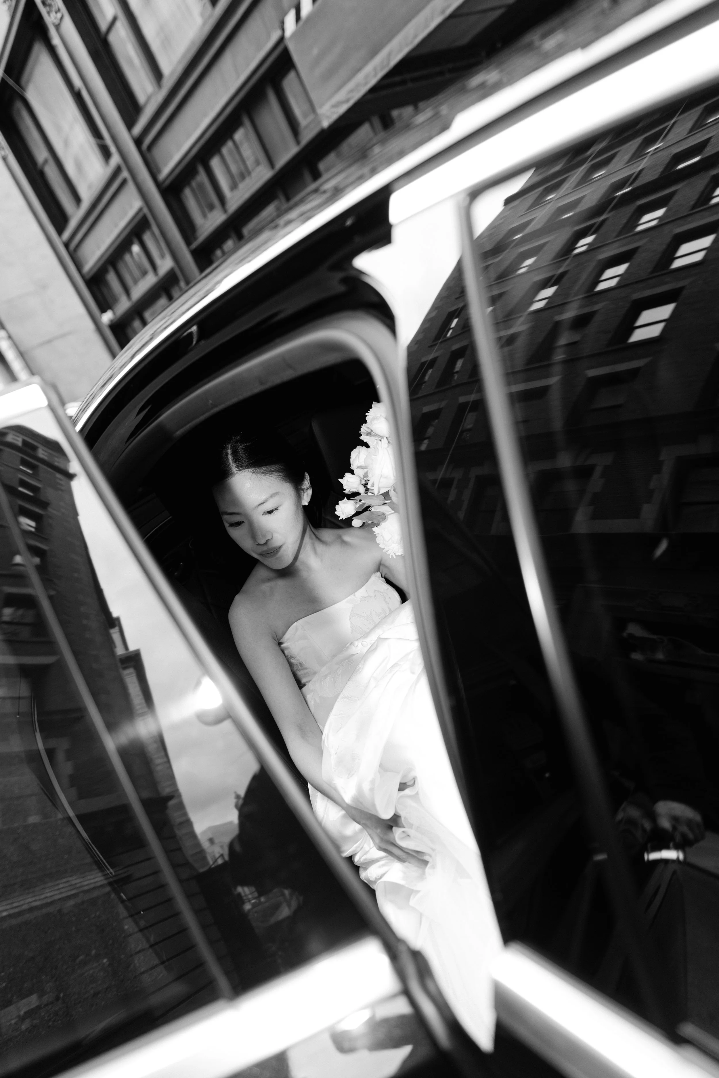 A woman in a wedding dress sitting inside a car, seen through the car's side mirror, with city buildings reflected in the mirror.