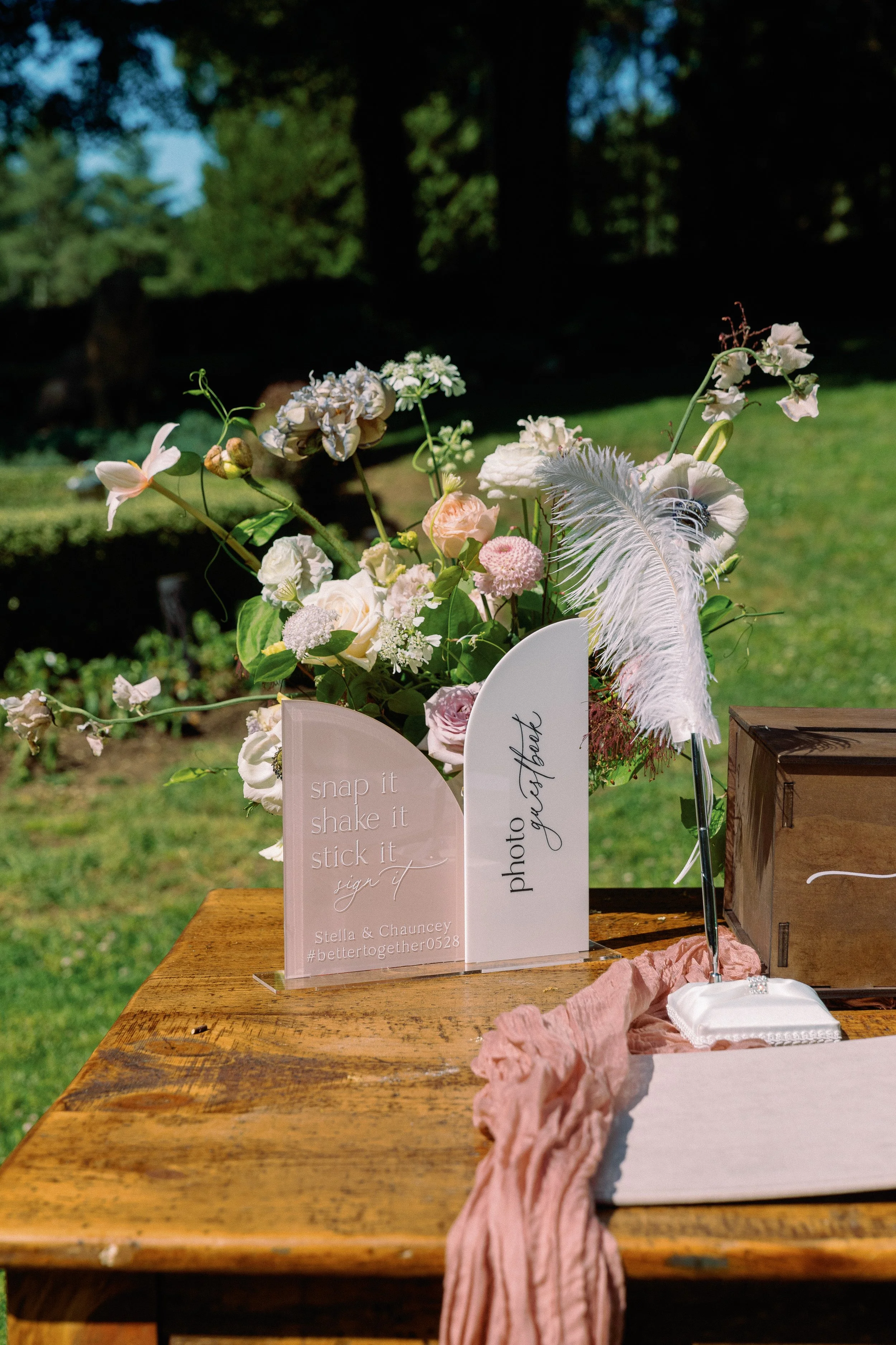 A wooden table holding decorative signs, a floral arrangement, and a ring display with a feathered pen for guest signatures at an outdoor wedding or event.