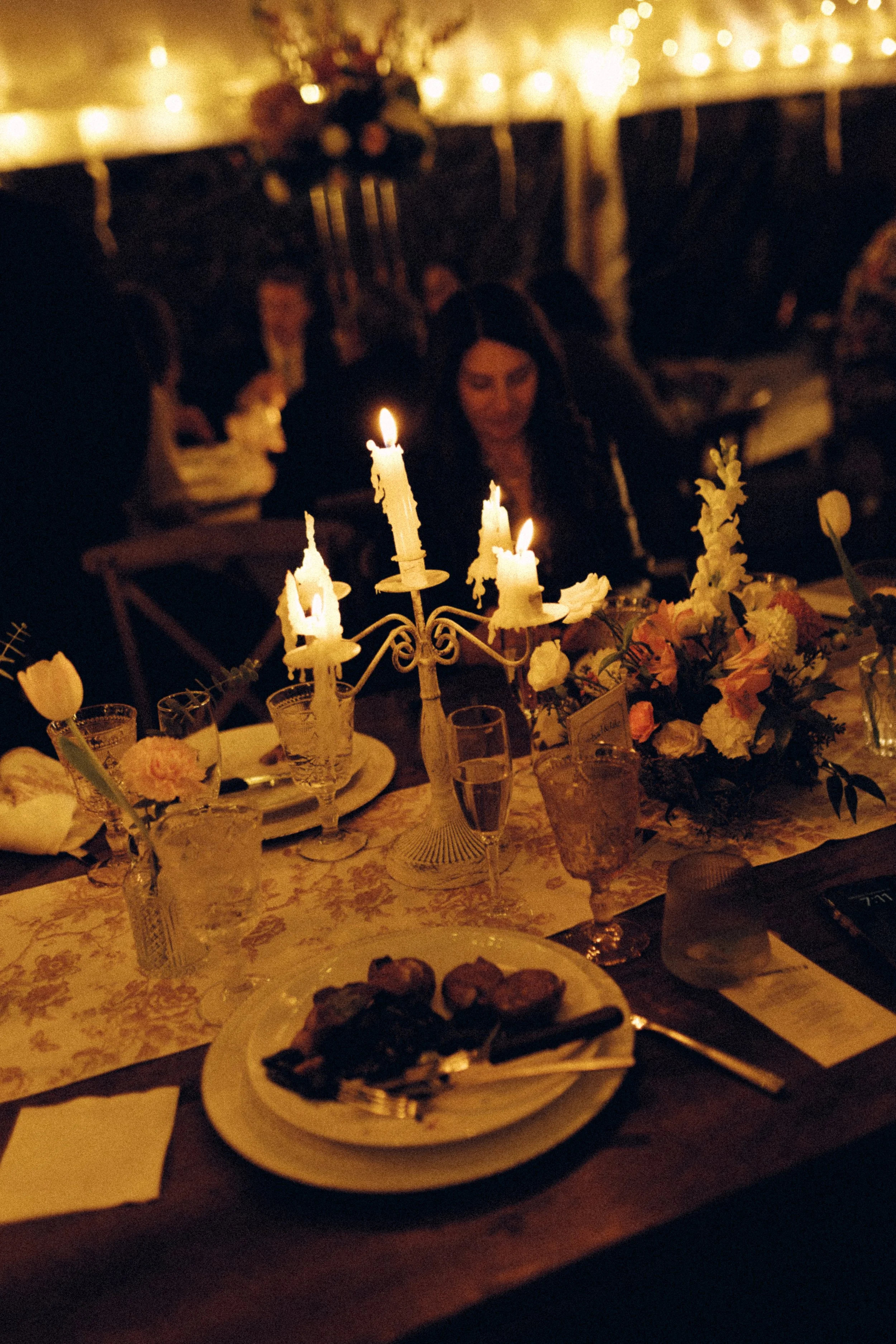 A dimly lit dinner table with a lit candelabra, floral arrangements, place settings, and a plate of food, with guests seated in the background.