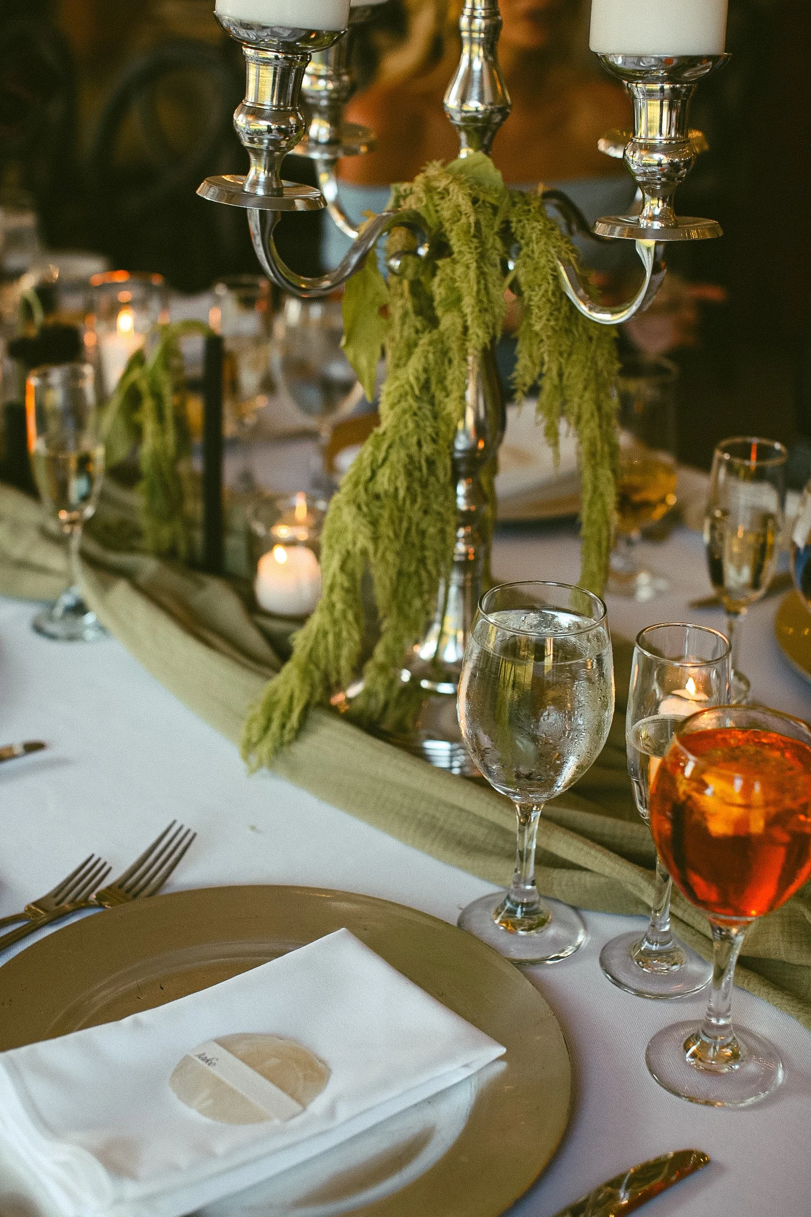 Elegant banquet table with silver candelabra, hanging greenery, wine glasses, a glass of orange-colored cocktail, and place settings with folded white napkins.