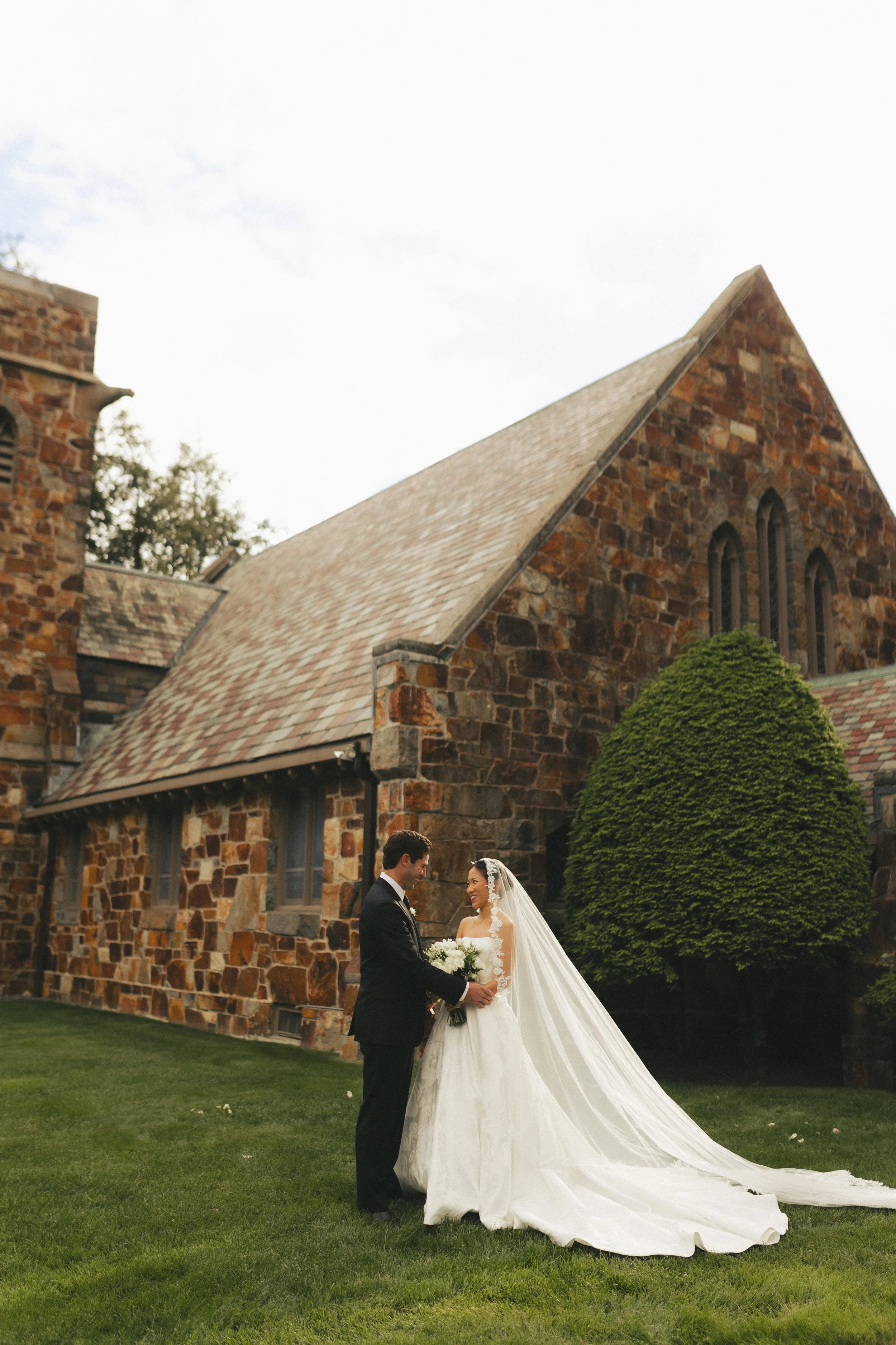 A bride and groom standing in front of a stone church, exchanging vows, with green grass and a bush nearby.