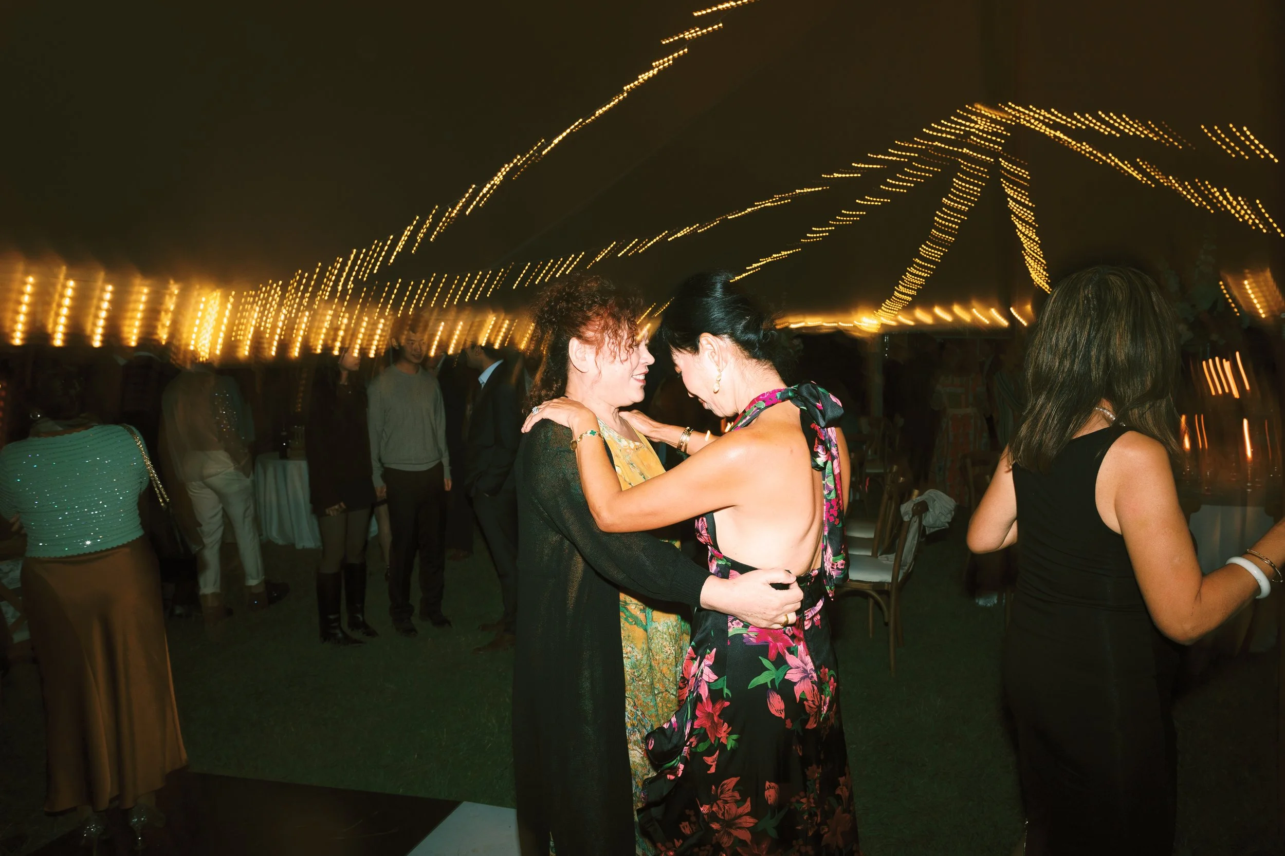 Two women dancing closely at a party under string lights with other guests in the background.