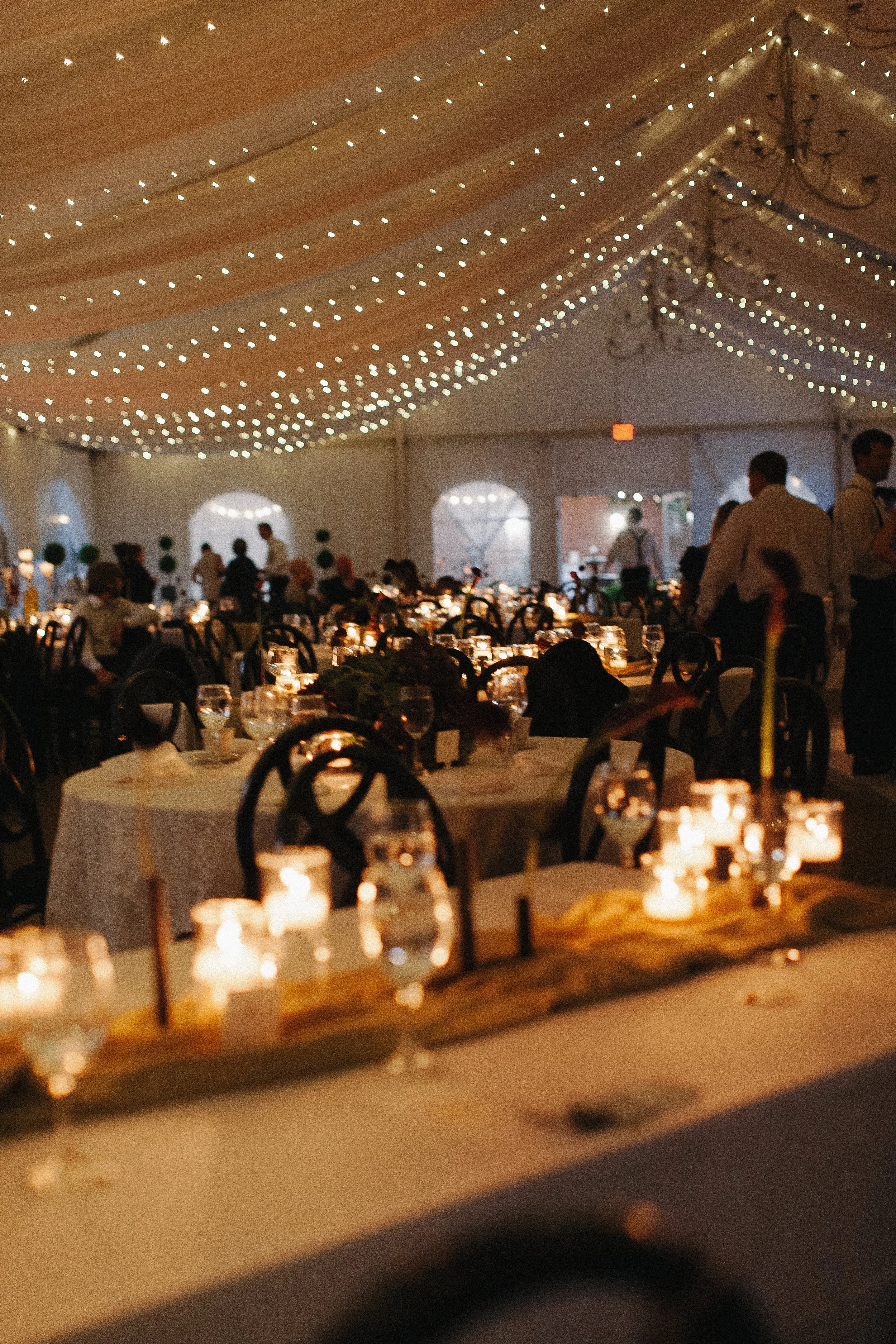 A dimly lit banquet hall decorated with string lights draped from the ceiling, with tables set with candles, glasses, and tableware for a formal event.