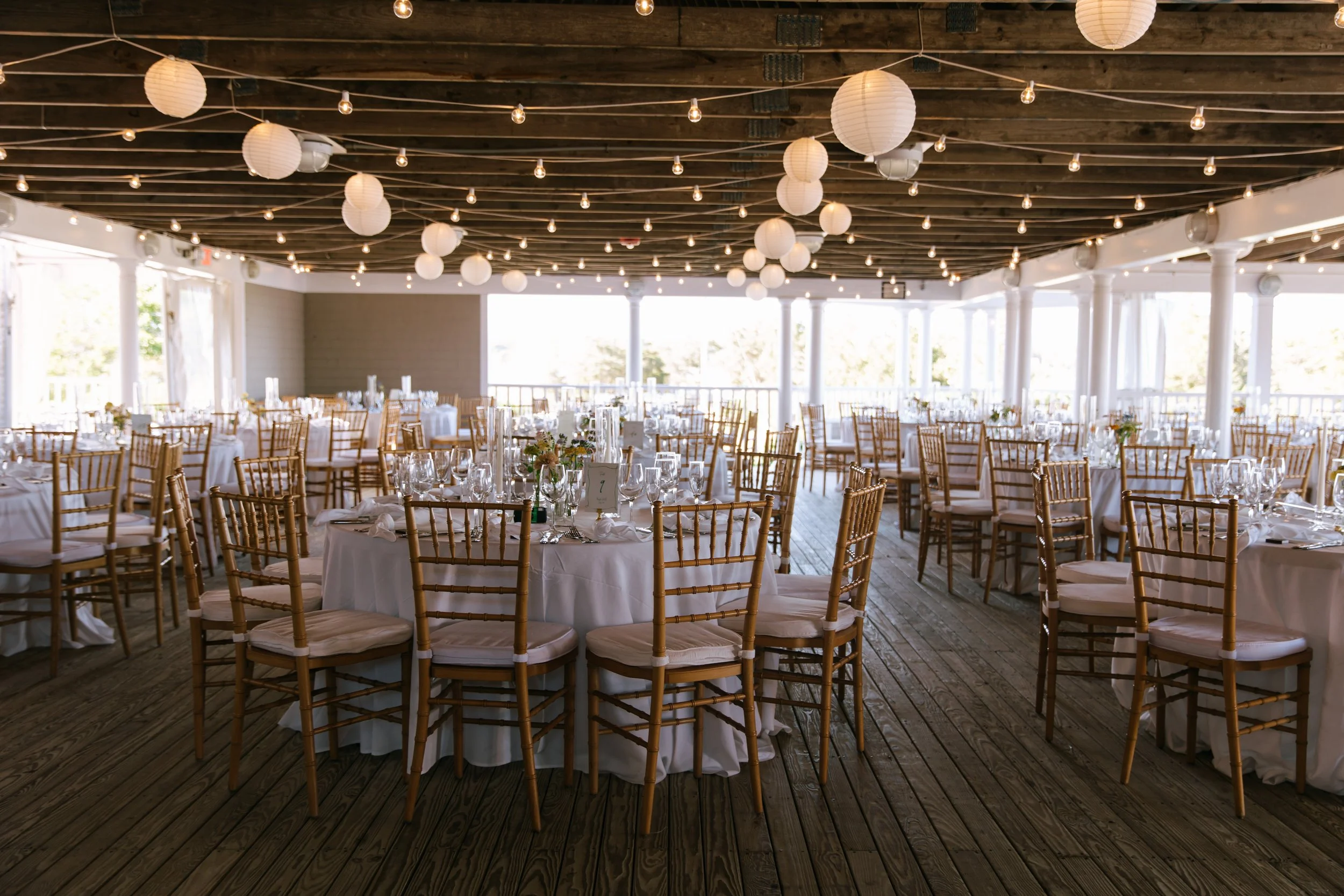 Indoor wedding reception decorated with white tablecloths, glassware, and flower centerpieces, with multiple round tables and gold-chair seating, illuminated by hanging paper lanterns and string lights.