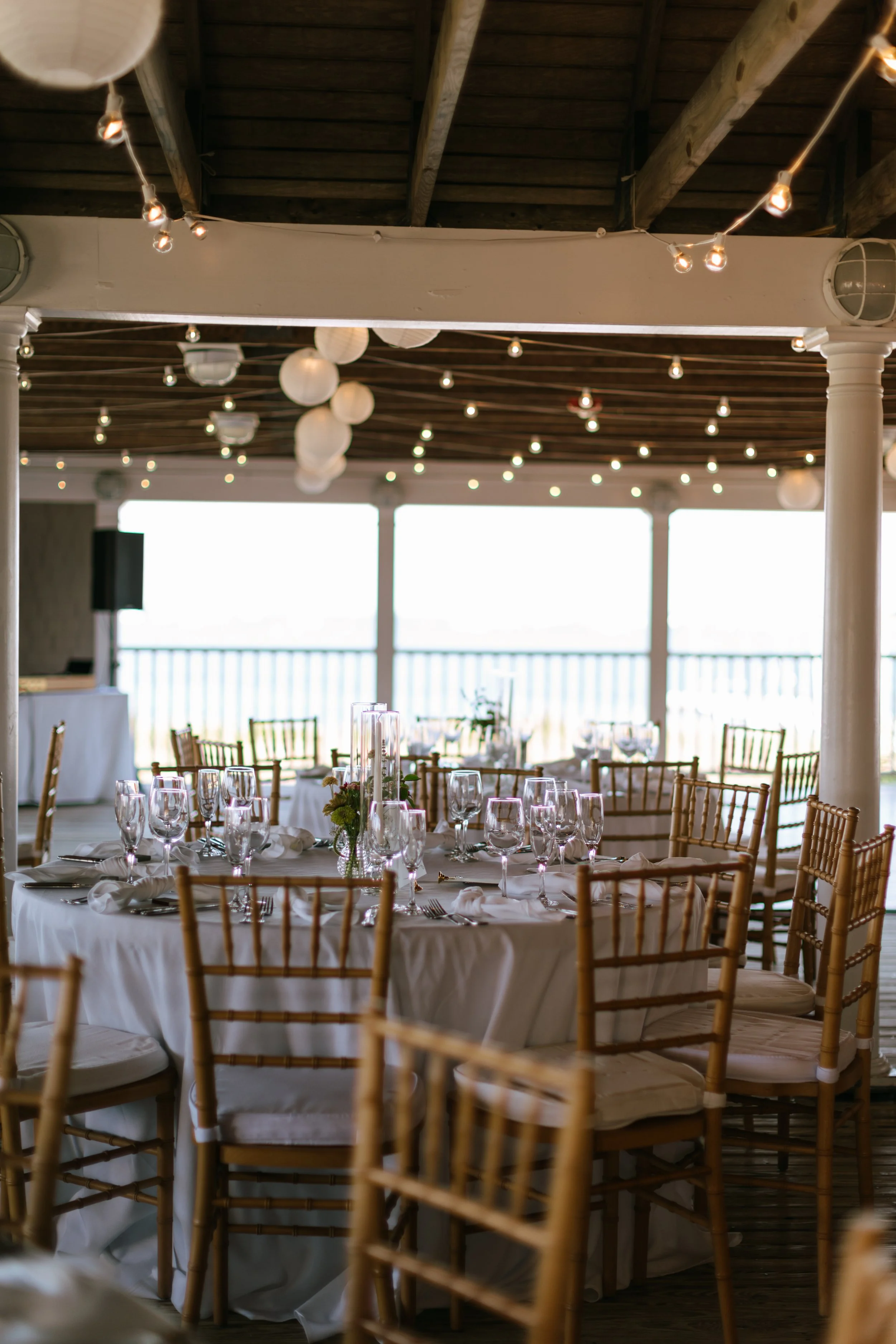 Decorated table at an event venue with wine glasses, napkins, silverware, and floral centerpieces, under string lights and paper lanterns, with a view of the outdoors through large windows.