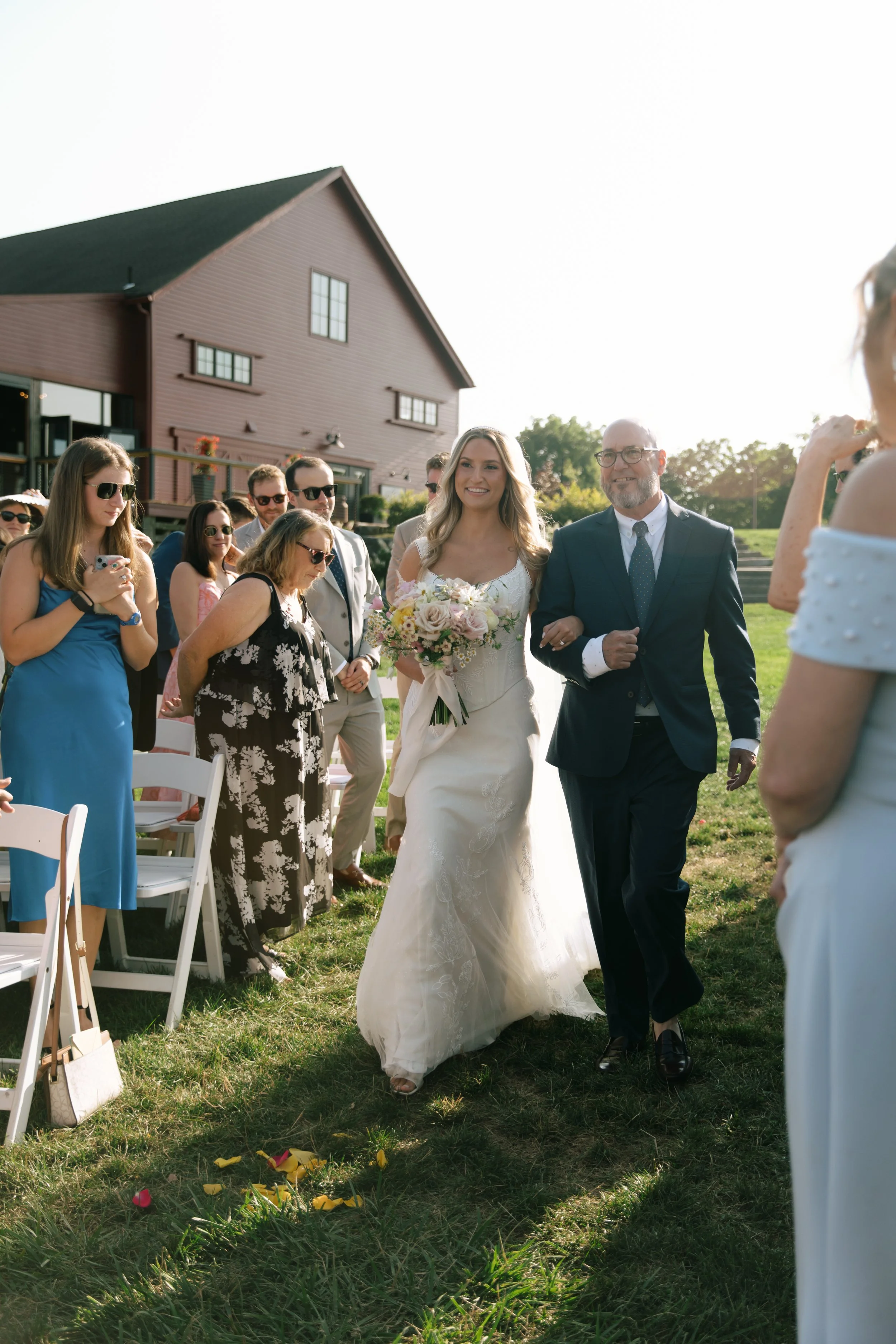 A bride walking down the aisle with her father at an outdoor wedding ceremony on a sunny day.