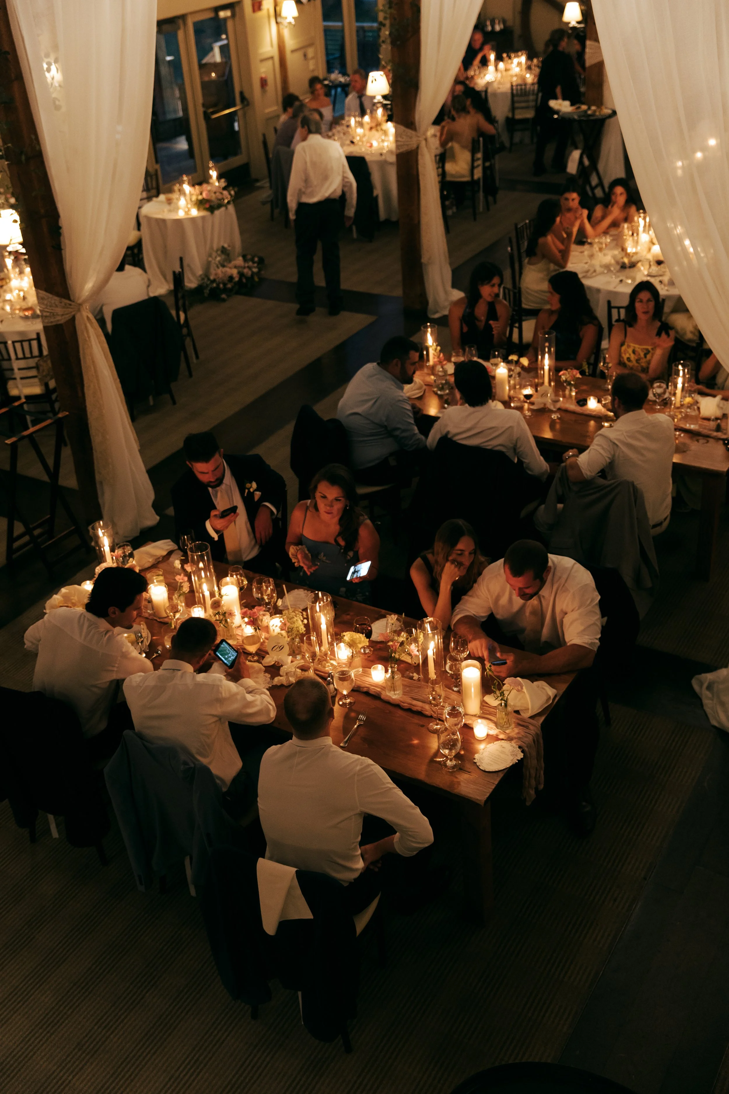 People dining at a wedding reception at Barn at Gibbet Hill Groton, sitting at long tables decorated with candles and flowers, in a warmly lit room with draped curtains. 