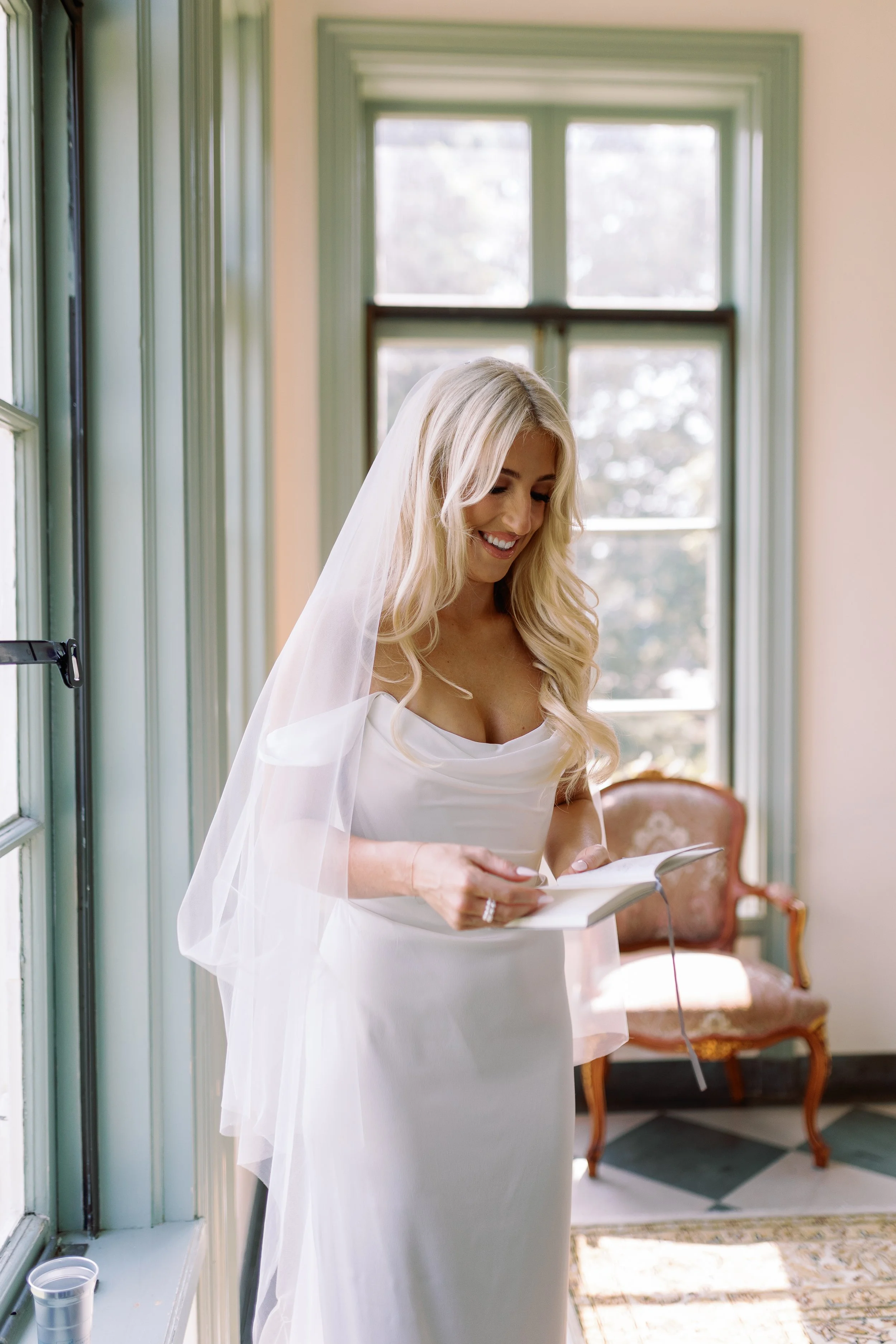 A bride in a white wedding dress and veil looks at a book, smiling, inside a room with large green-framed windows and vintage furniture.
