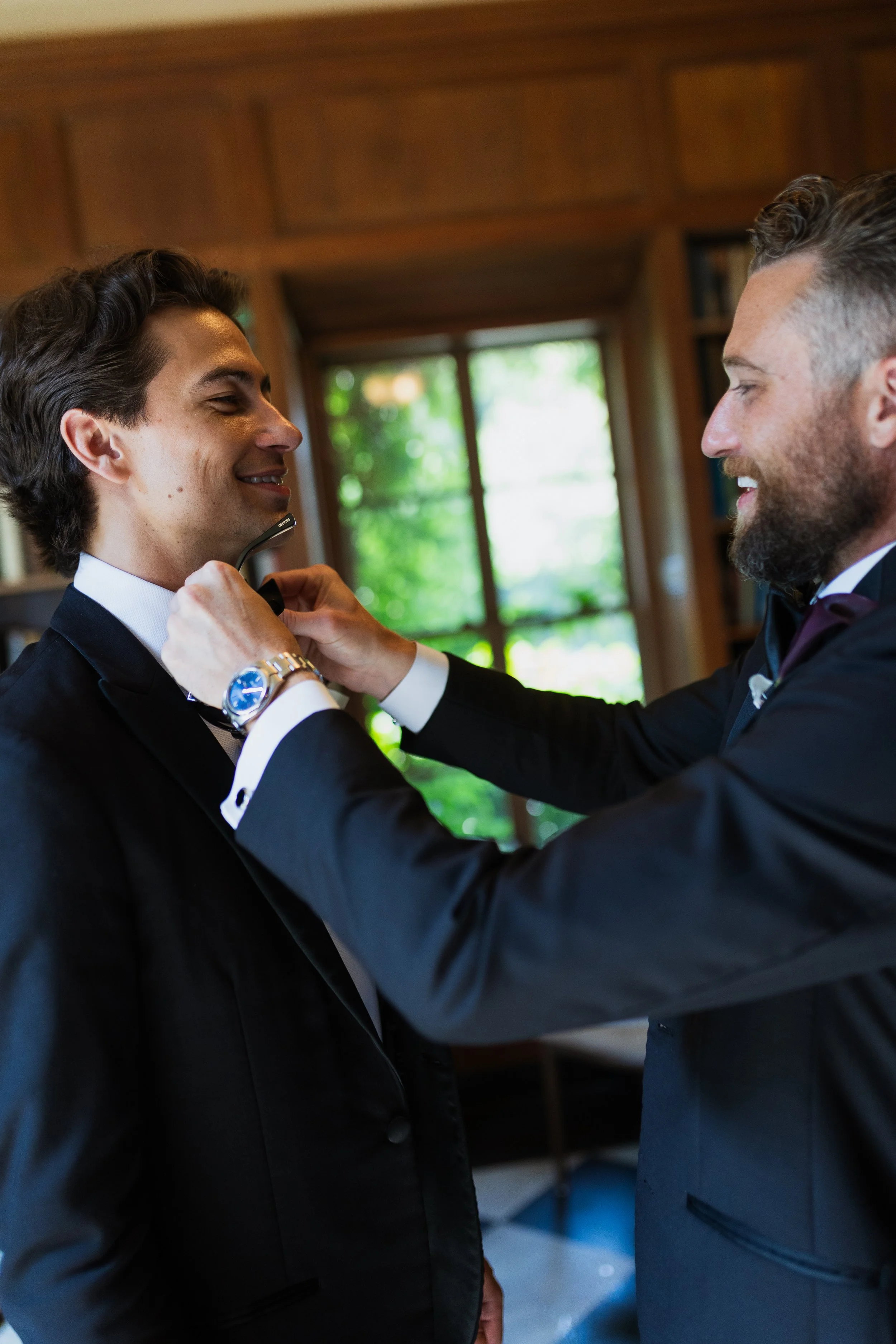 Two men in tuxedos preparing for a formal event, one adjusting the other's bow tie in a well-lit room with large windows and greenery outside.