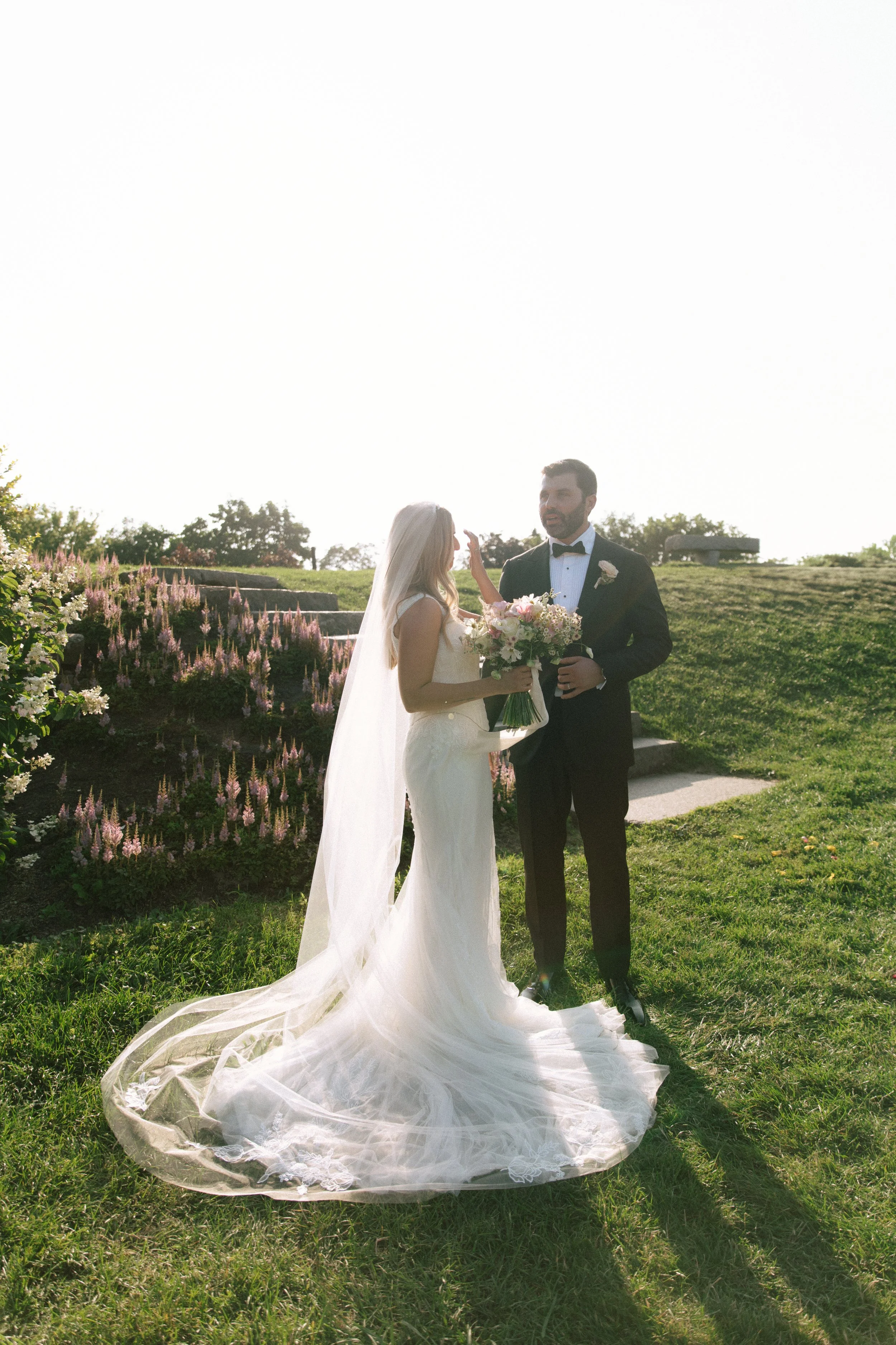 Bride and groom standing outdoors on a grassy area with stairs and flowering bushes, wedding ceremony scene, sun is bright behind them.
