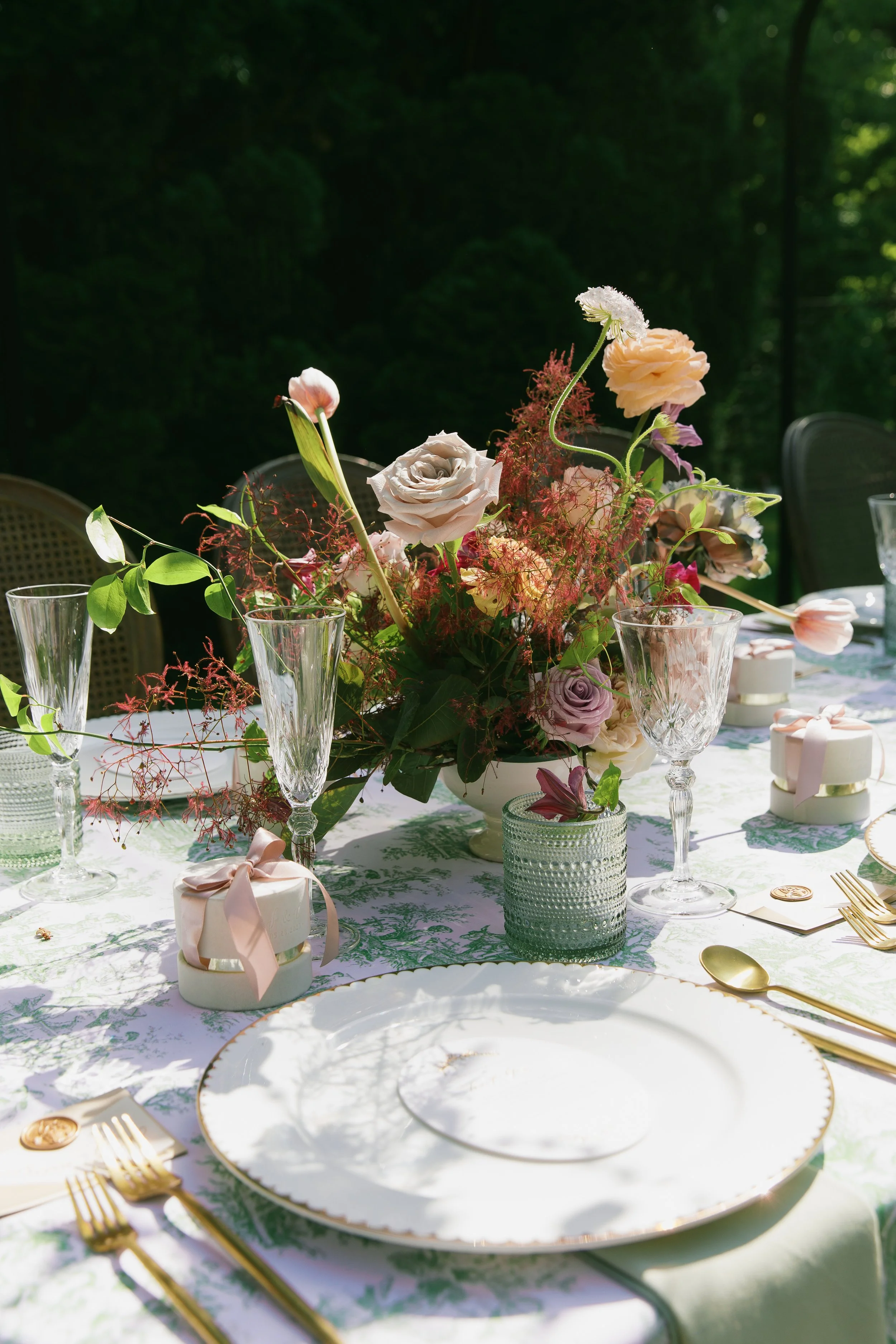 Elegant outdoor table setting with a floral centerpiece, white plate with gold trim, gold utensils, pink favor boxes, and gold-rimmed glasses in a lush green outdoor setting.