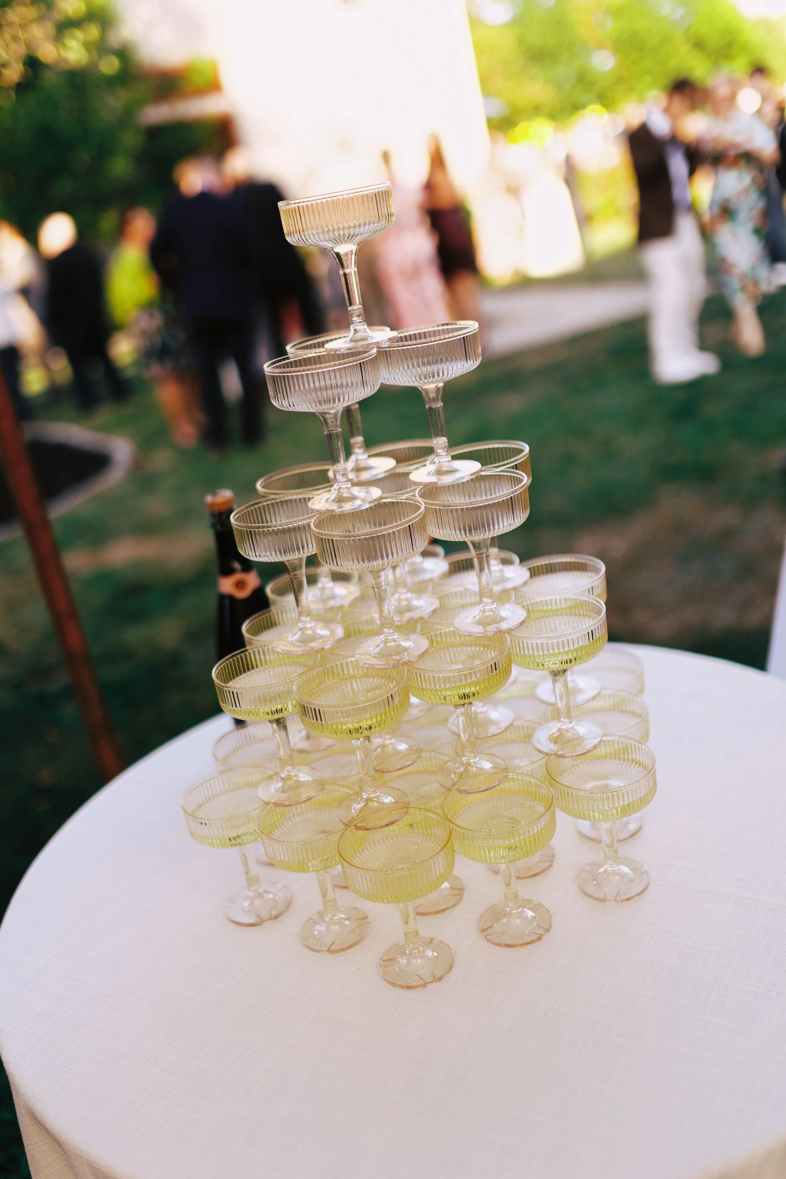 A pyramid of champagne glasses with green liquid on a round table at an outdoor event.