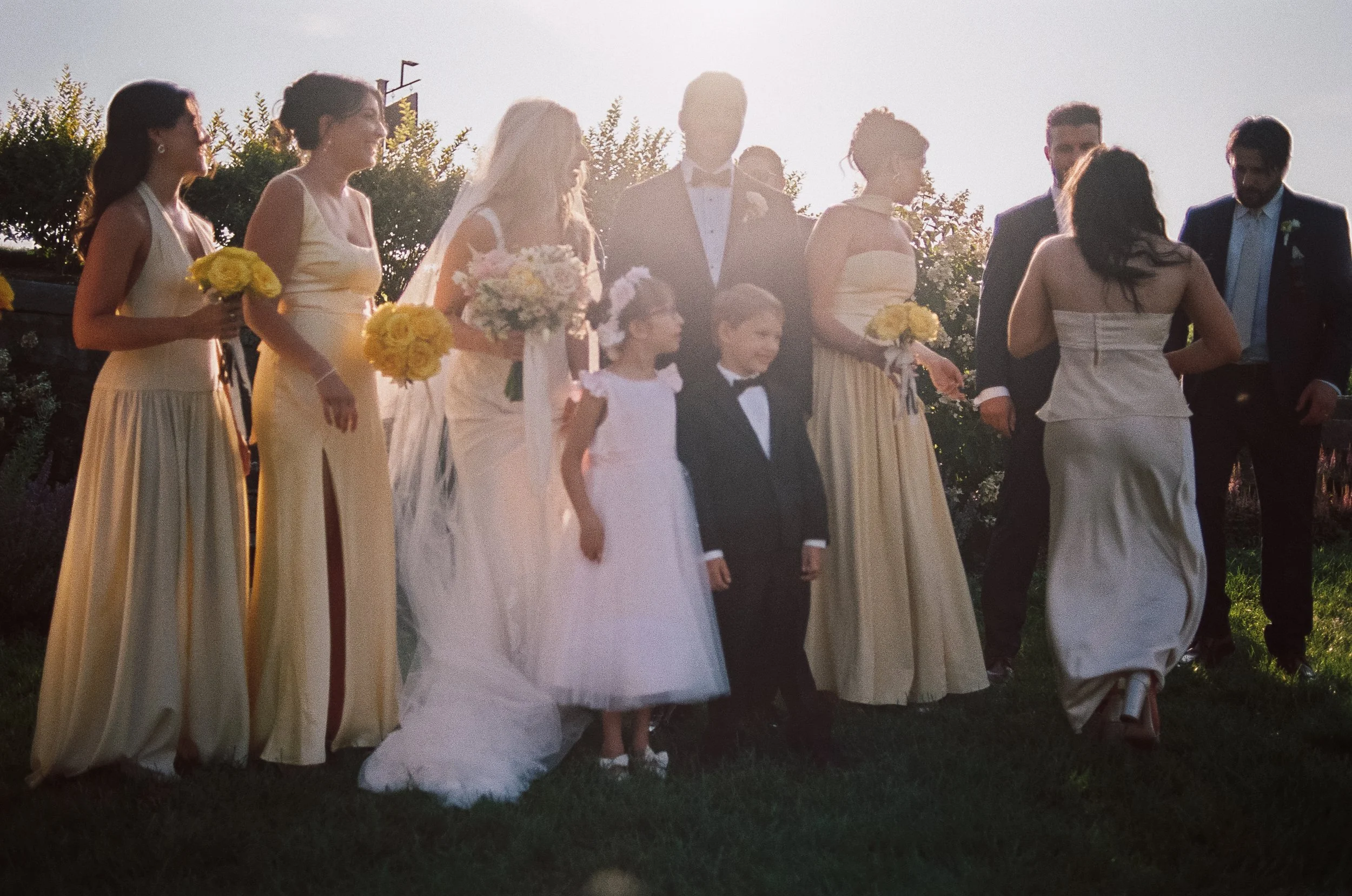 A group of wedding guests outdoors during sunset, including a bride in a white gown with a bouquet, a groom in a tuxedo, children, and women in matching yellow dresses holding yellow bouquets, standing on grass with greenery in the background.