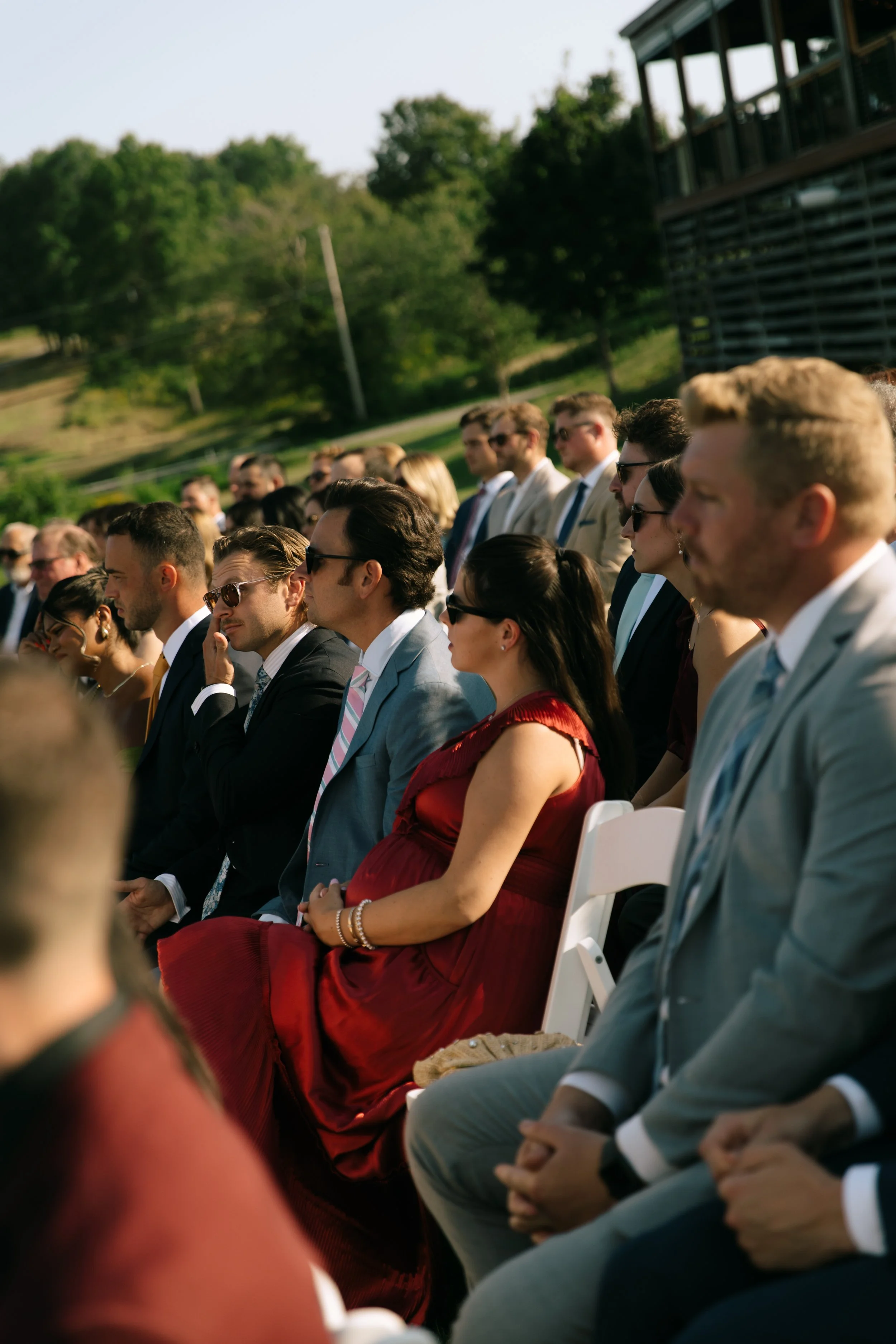 People attending an outdoor event, sitting on white chairs, dressed in formal attire, on a sunny day with green trees in the background.