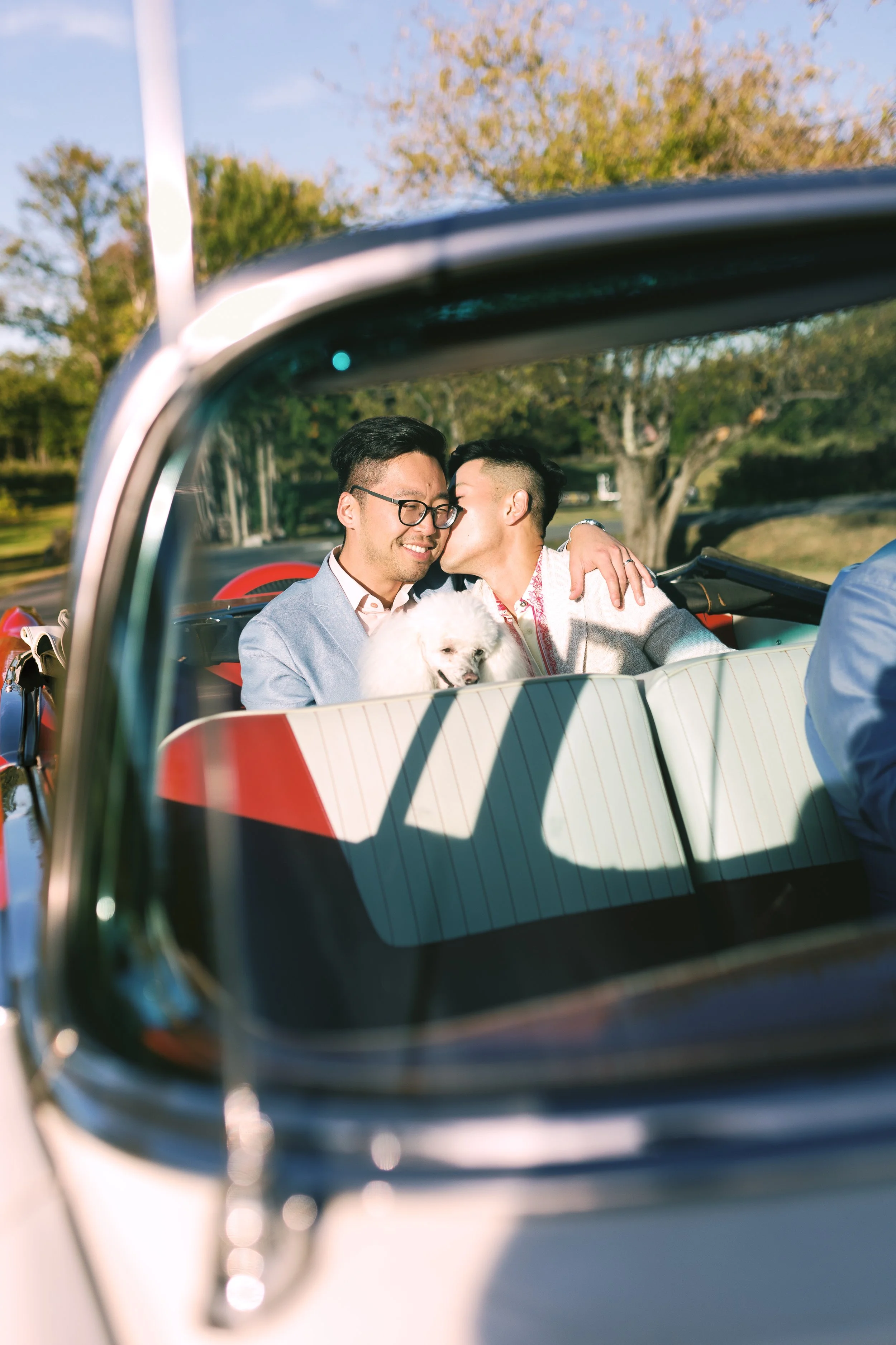 Two men sitting in a vintage convertible car, one kissing the other on the cheek, with a small white dog between them, in a park setting during daytime.