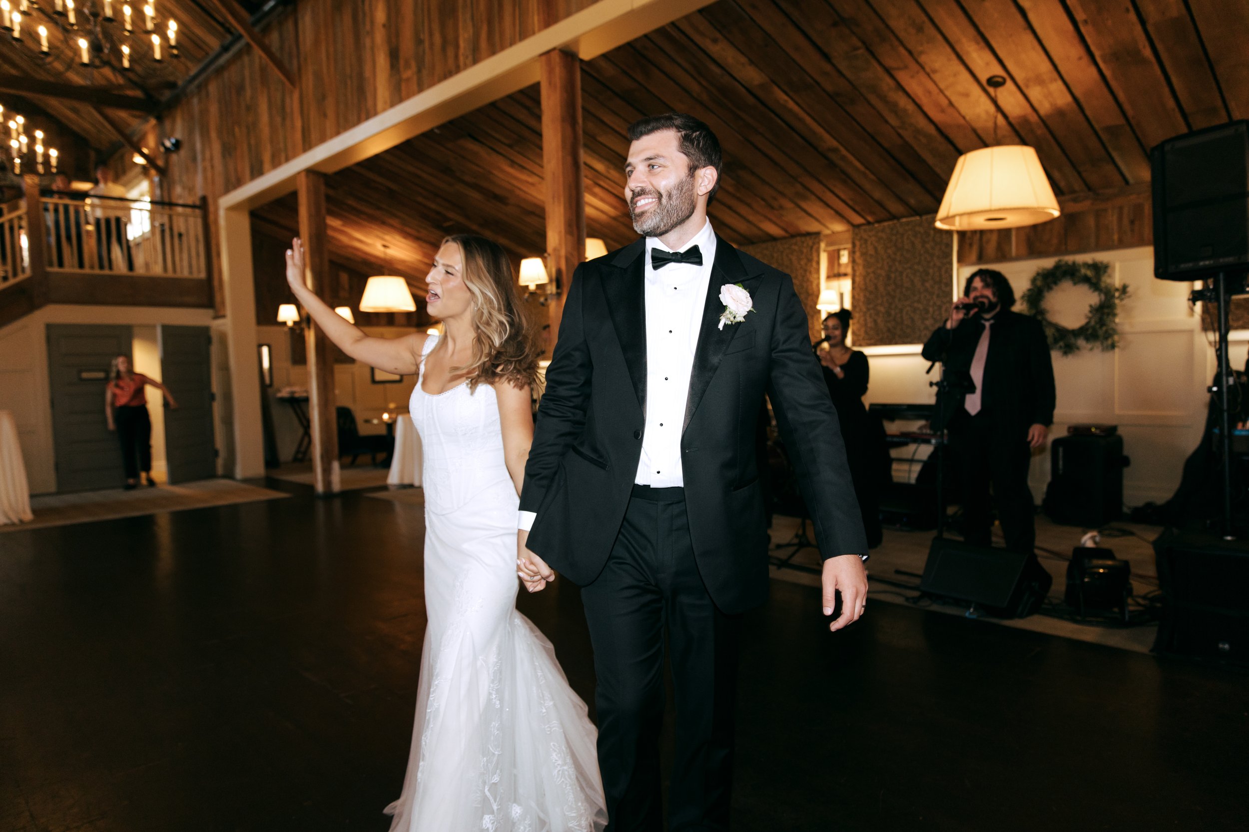 Bride and groom holding hands and dancing at their wedding reception in a rustic venue, with live band playing in the background.