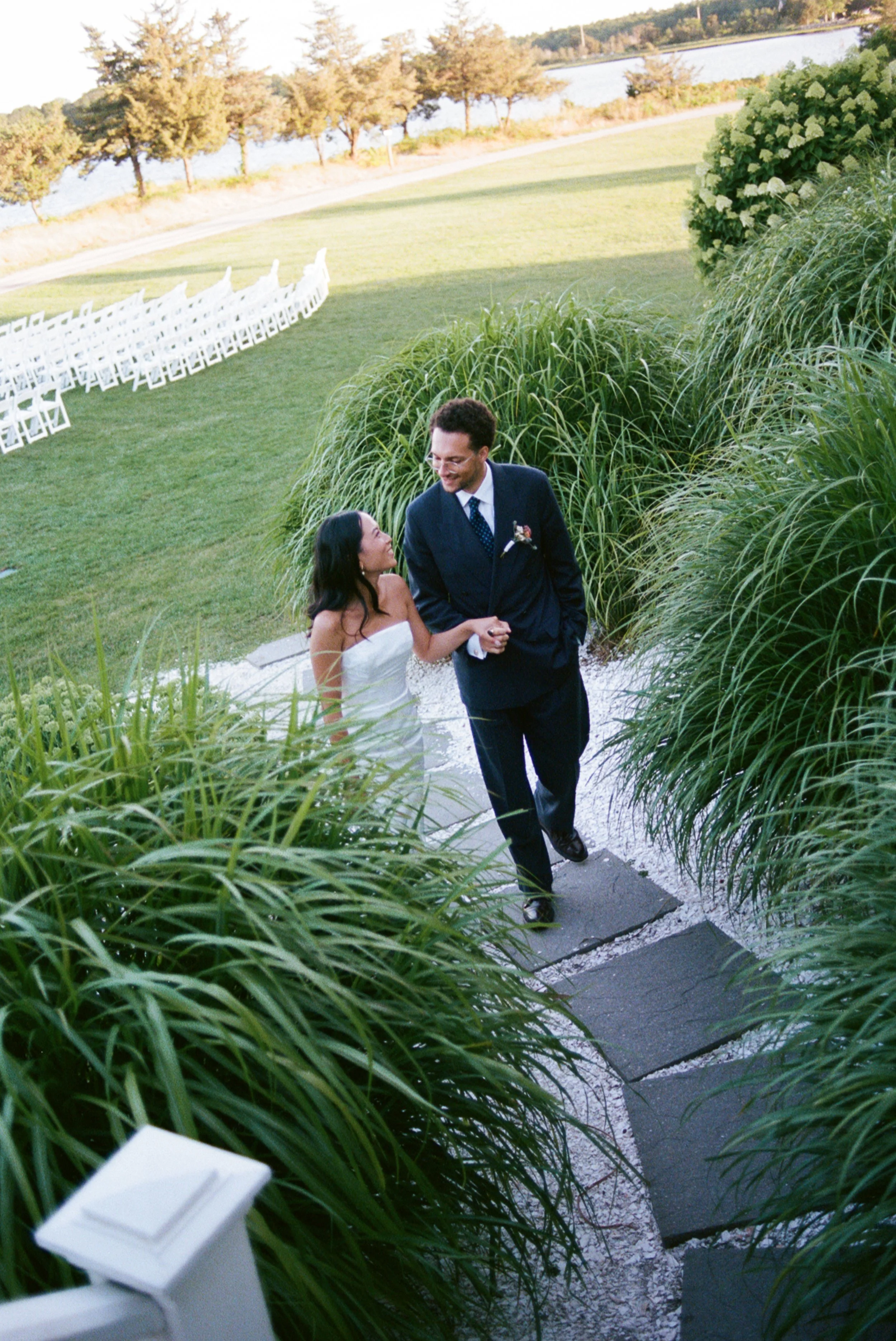 A newlywed couple walking hand in hand on a stone path surrounded by lush green bushes, with white chairs arranged outside in the background near a body of water.