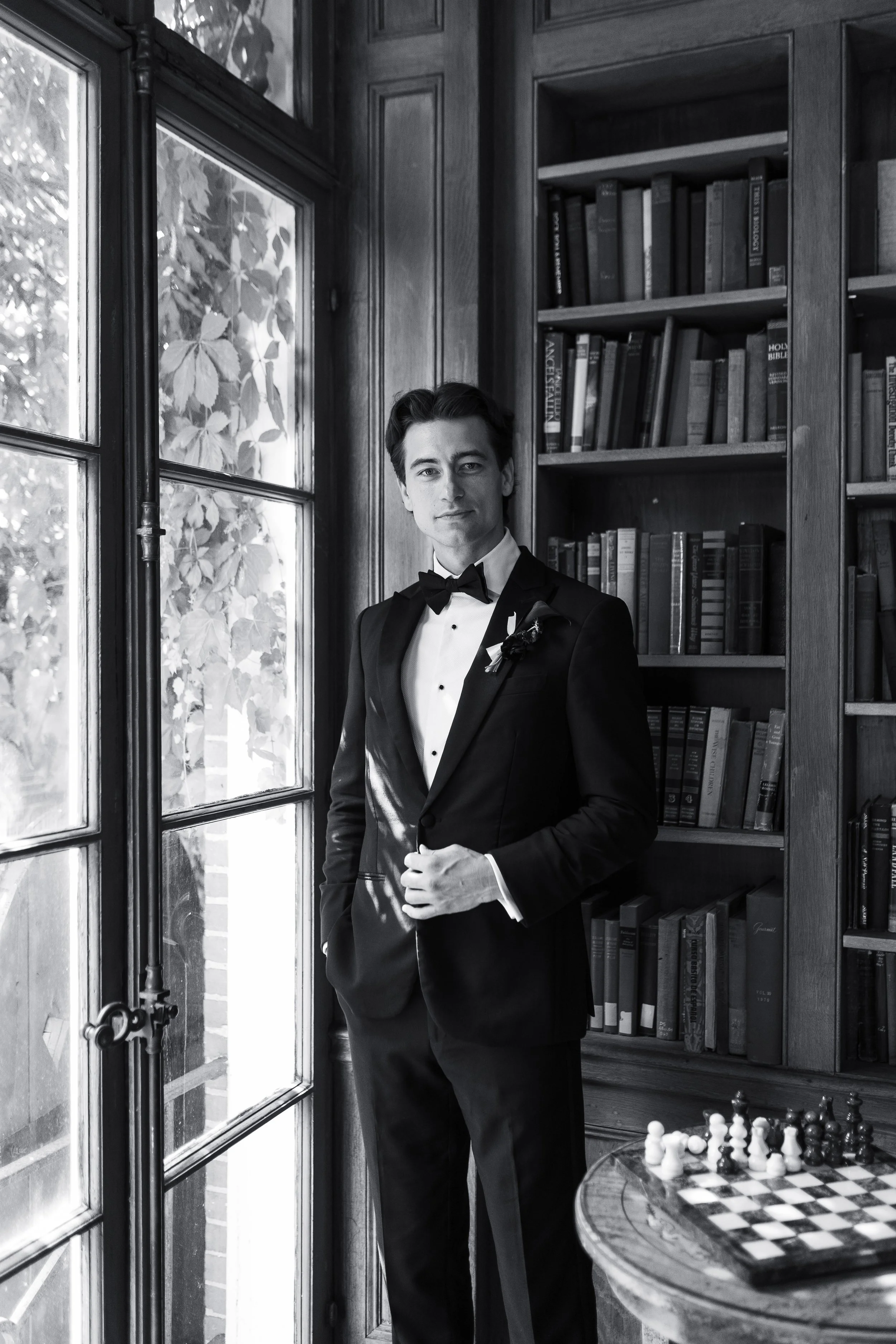 A young man in a black tuxedo with a bow tie and boutonnière stands indoors near a large window with a wooden bookshelf in the background. A chessboard is on a small table in the foreground.