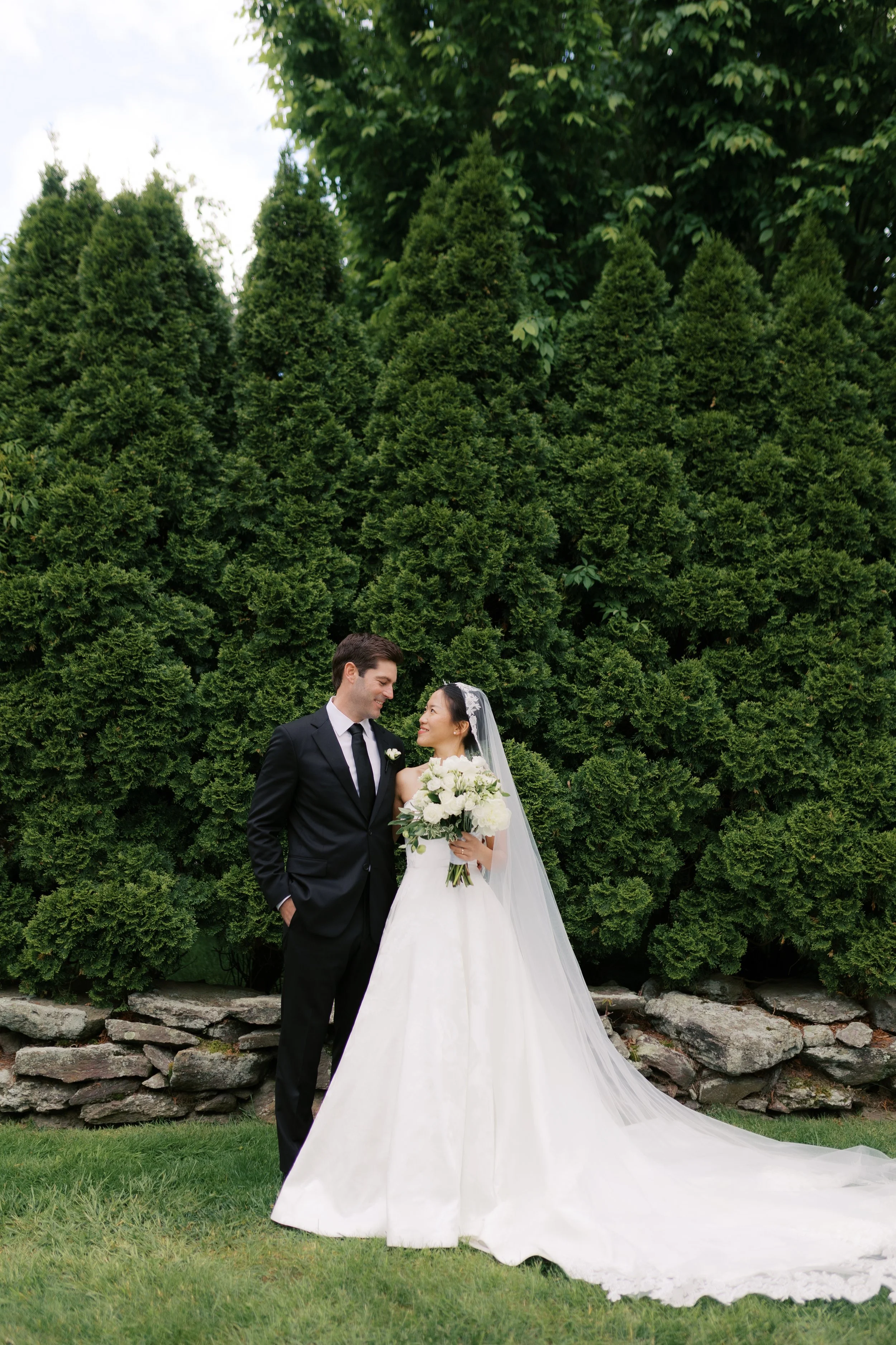 A bride and groom standing outdoors in front of green bushes, with the bride holding a bouquet of white flowers and wearing a wedding dress with a long veil, while the groom wears a black suit and tie, and they look at each other affectionately.