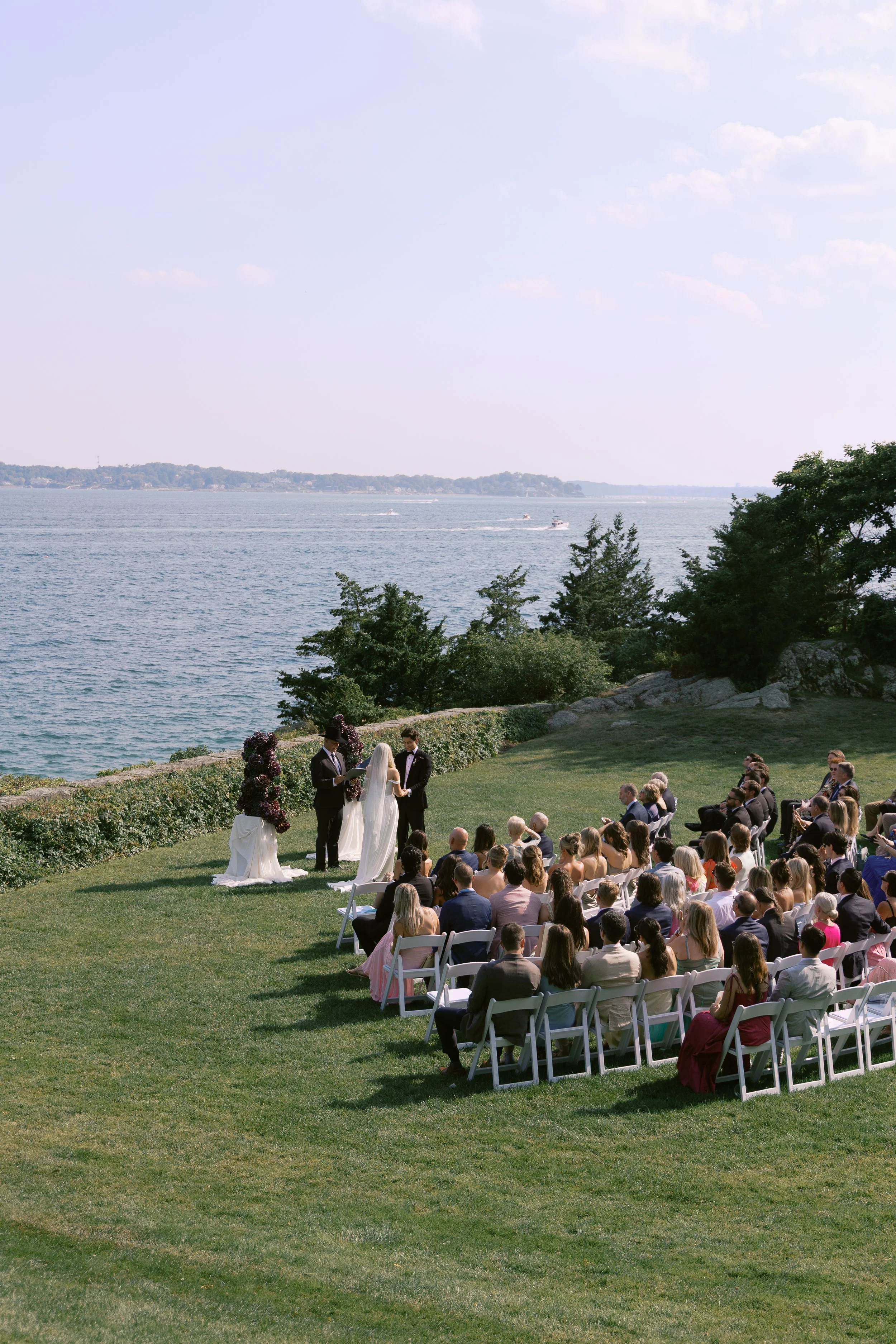 An outdoor wedding ceremony by a body of water with chairs and guests, a bride and groom, and officiant.