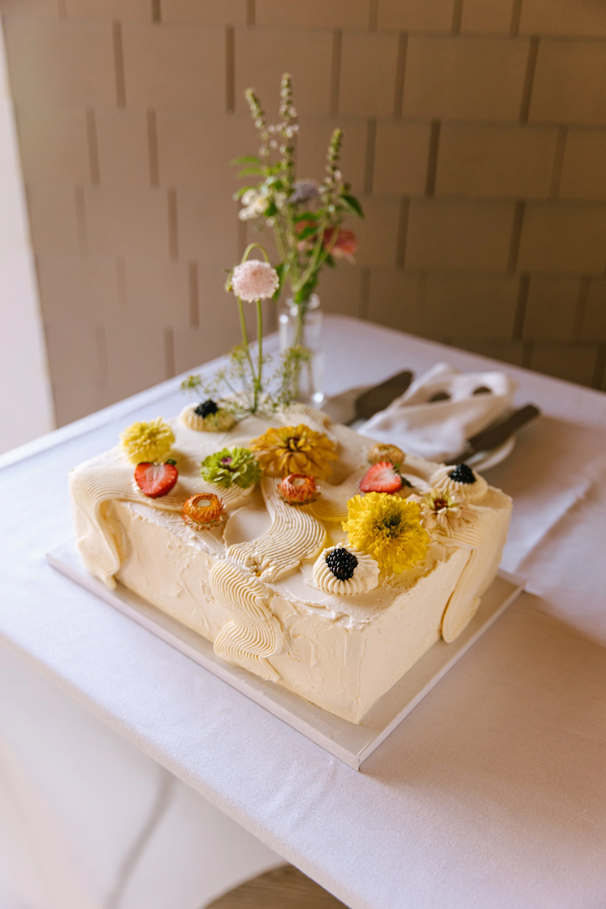 A square white cake decorated with flowers, strawberries, blackberries, and cream piping, placed on a white table with a floral arrangement in a vase in the background.