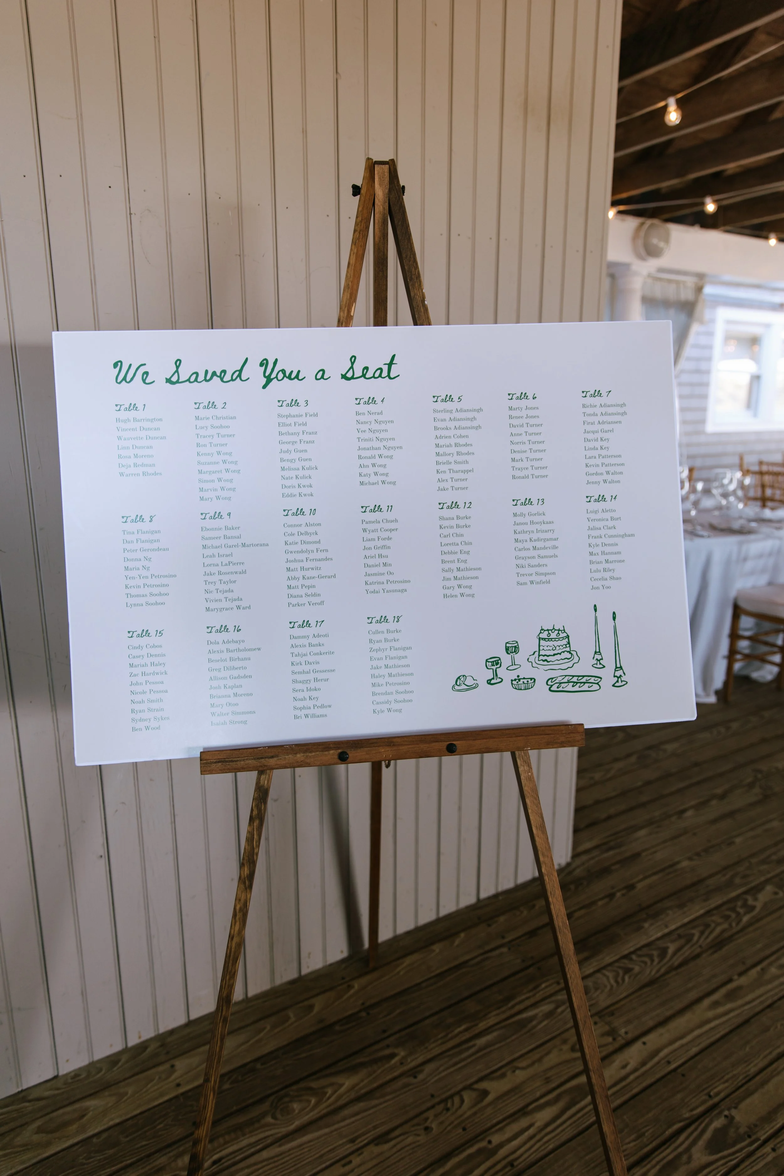 Wedding reception seating chart on a white board with green text, listing guest names assigned to tables, and simple green line illustrations of cakes, drinks, and utensils at the bottom.