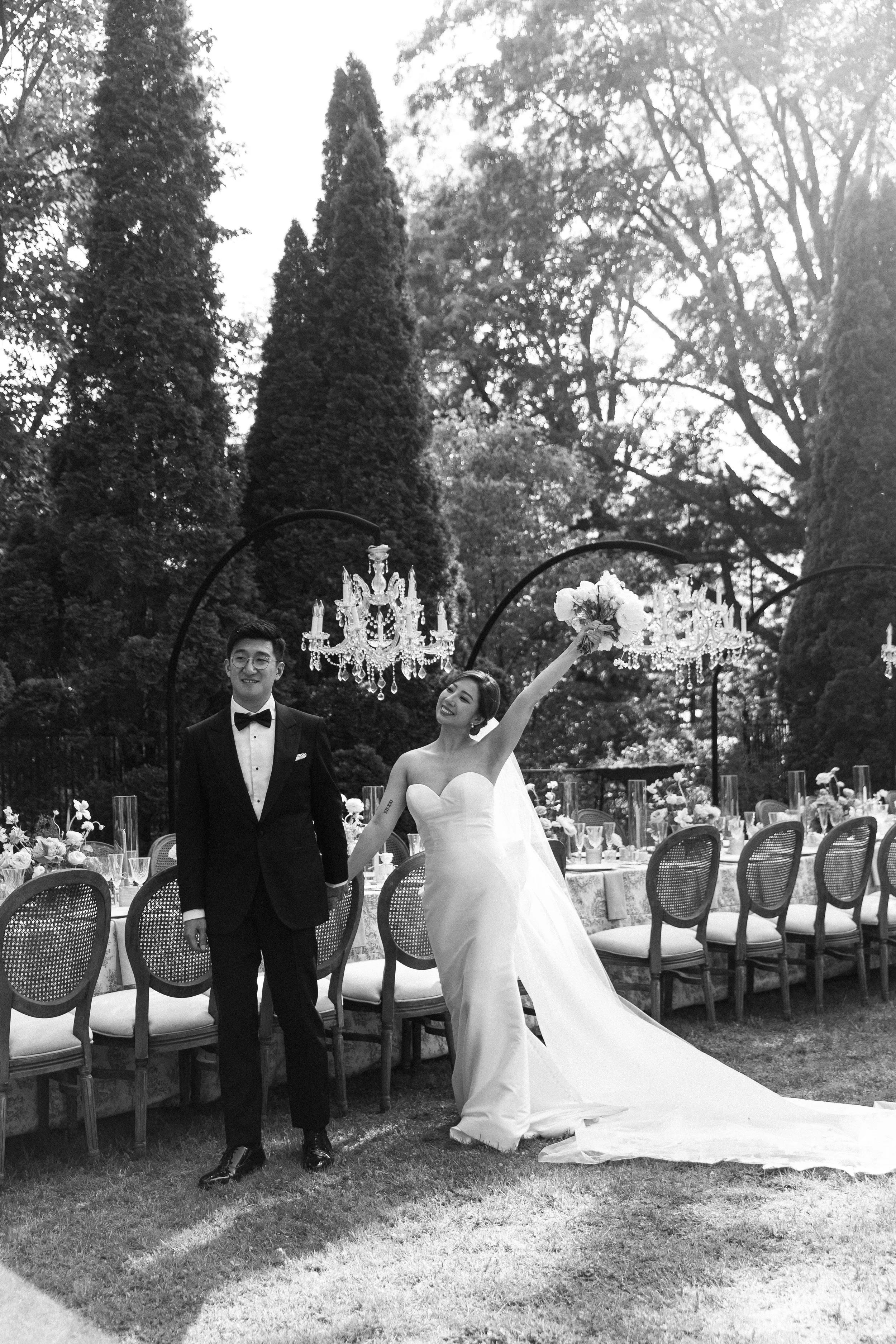 A bride and groom stand outdoors at a wedding reception, with the bride in a strapless wedding gown holding a bouquet, and the groom in a tuxedo; they are smiling and holding hands, surrounded by decorated tables and chandeliers in a garden setting.