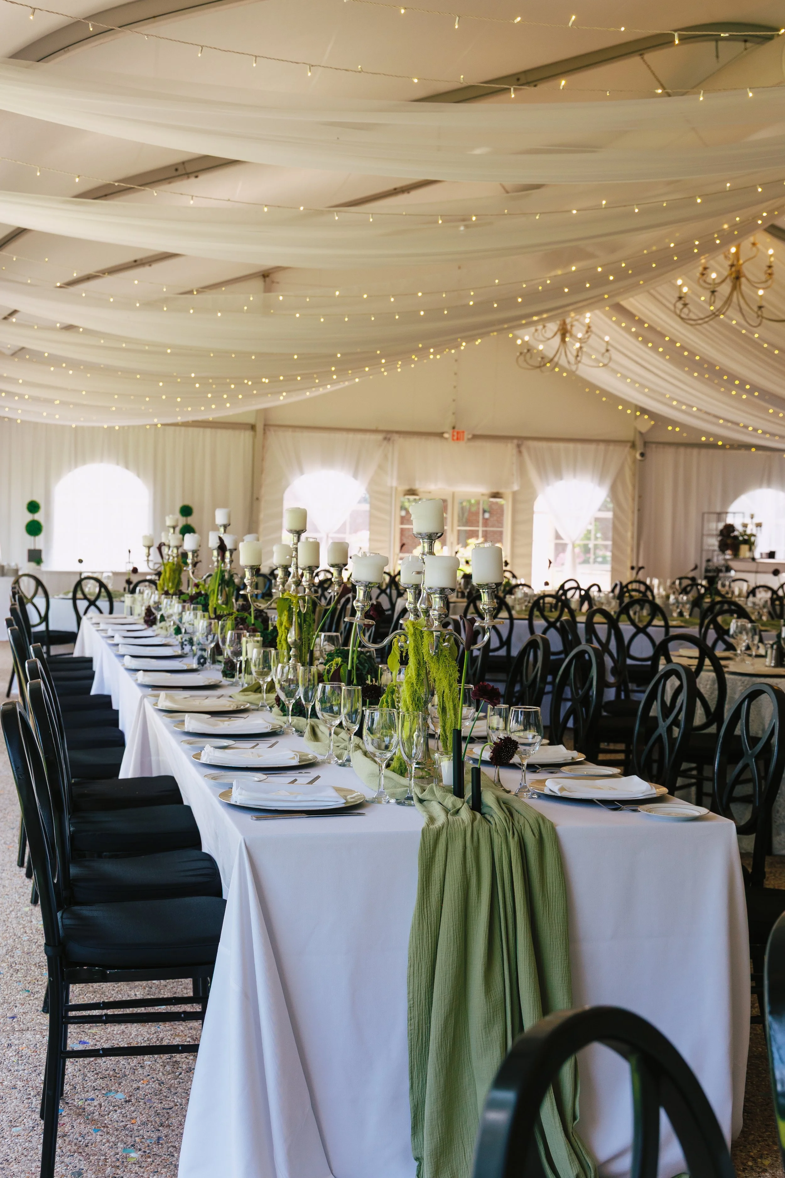Elegant banquet hall decorated for a wedding or special event with long tables, black chairs, candle centerpieces, and draped ceiling with string lights.