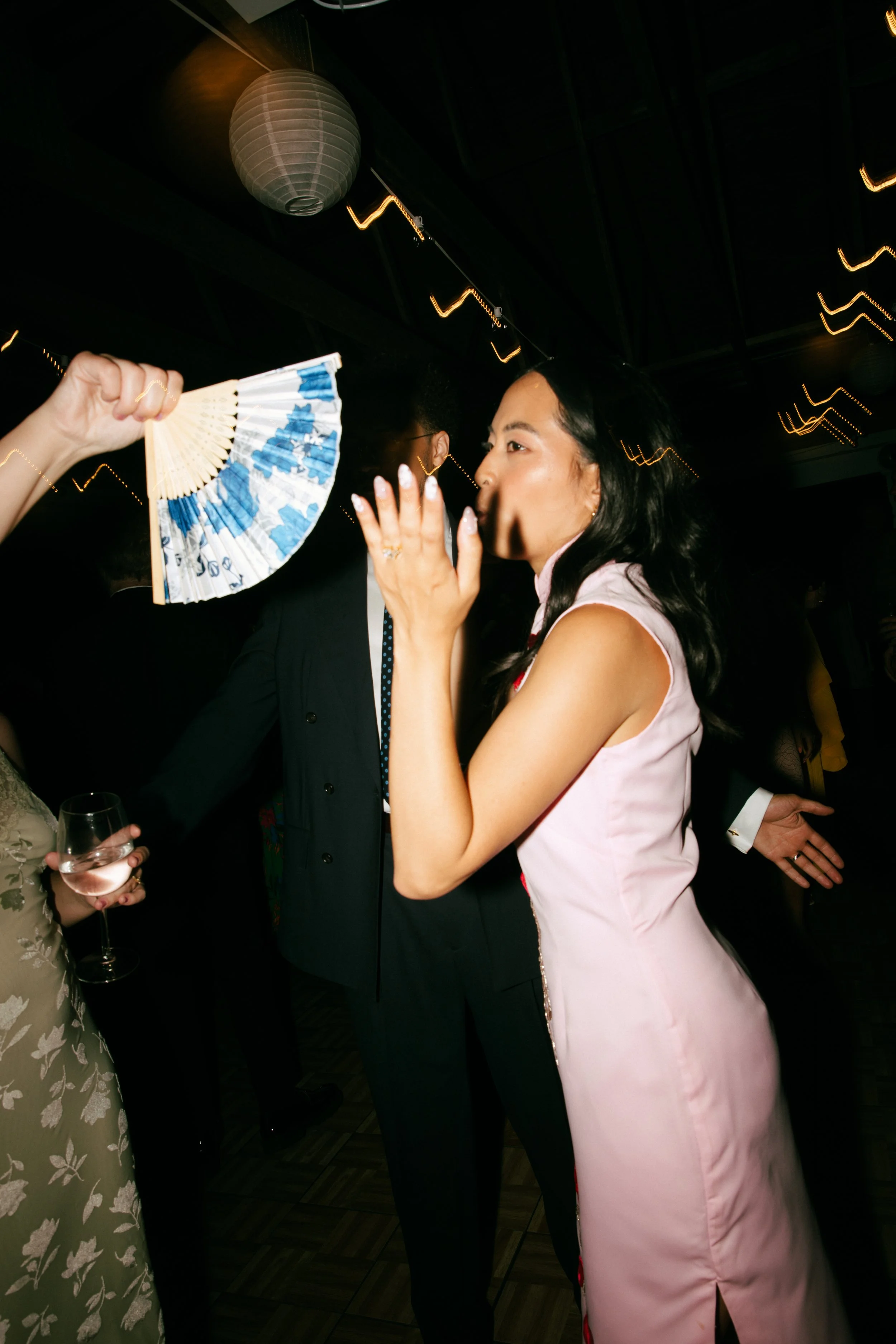 A woman in a pink dress whispering to a man at a party or celebration, with another woman holding a wine glass nearby and a person waving a decorative fan, in a dimly lit venue