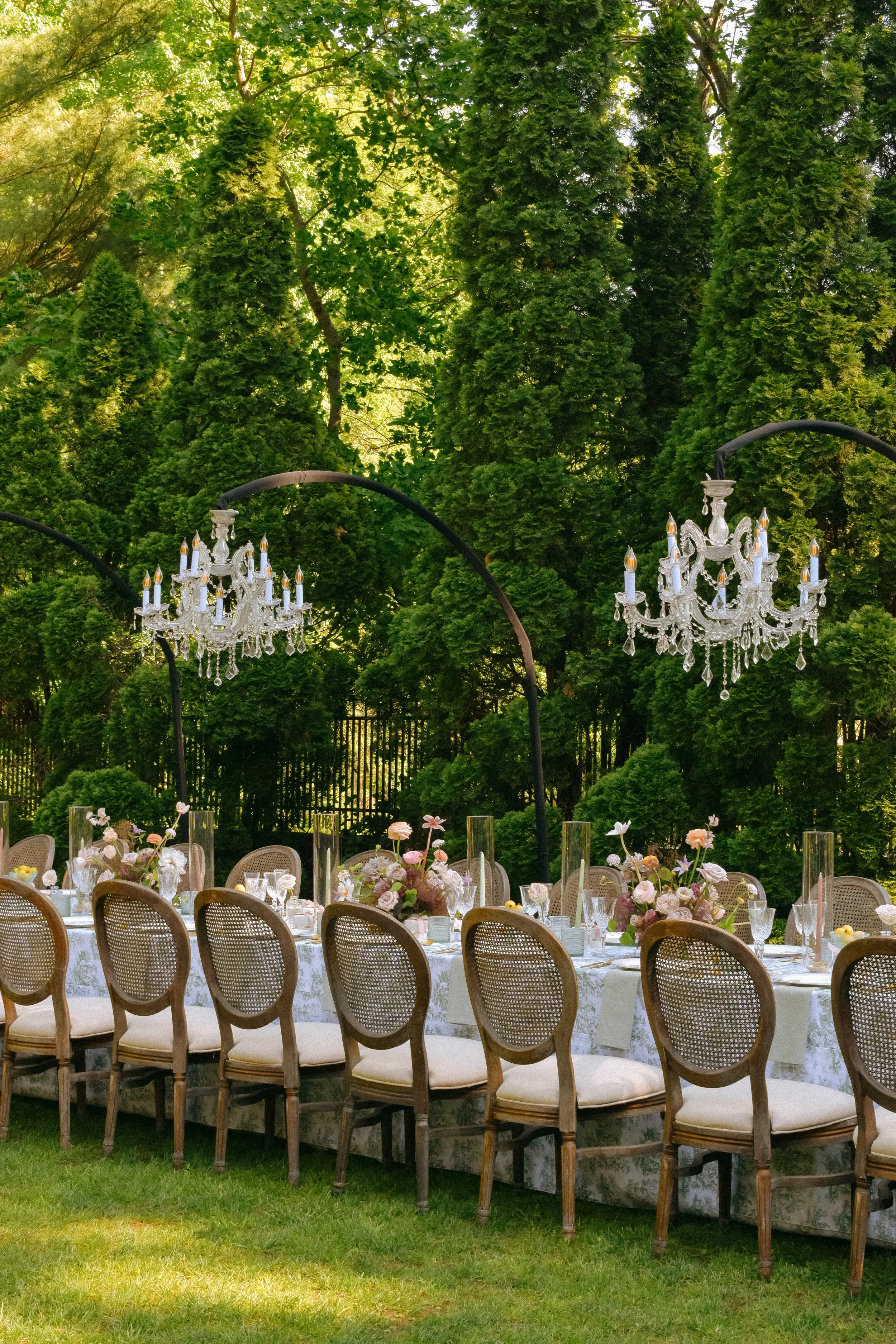 Elegant outdoor dining setup with a long table, floral centerpieces, chandeliers hanging above, and surrounded by lush green trees.