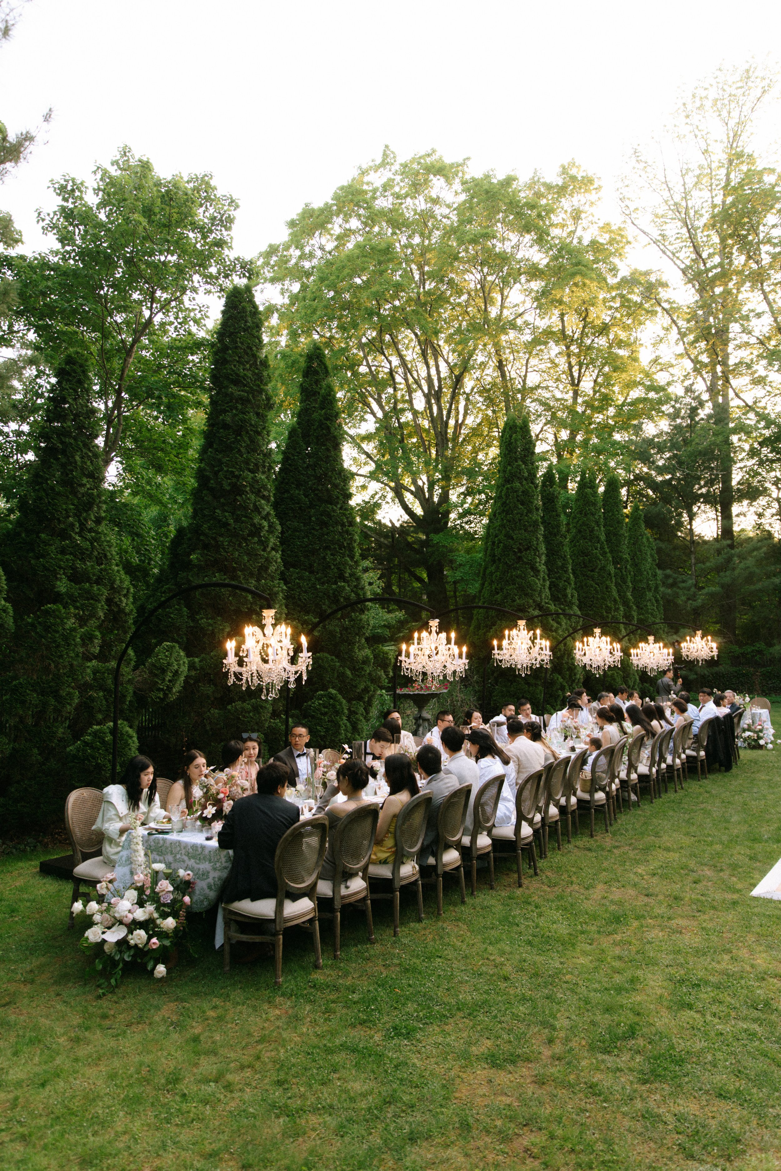 An outdoor dinner party with long table, elegant chandelier lighting, surrounded by green trees and landscaping.