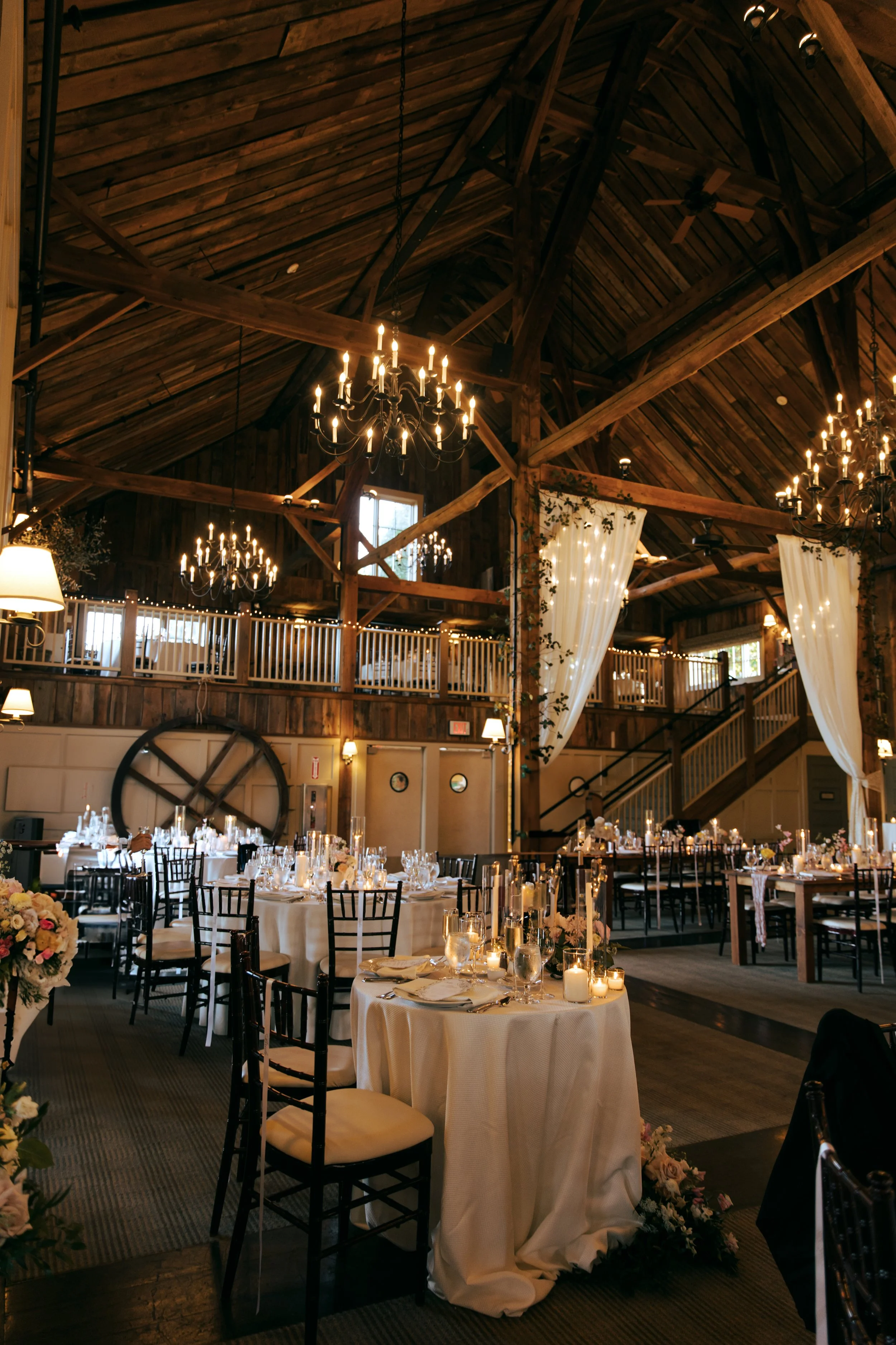 Interior of a rustic Barn at Gibbet Hill Groton wedding reception hall with wooden beams, chandeliers, and decorated tables with candles and floral centerpieces.