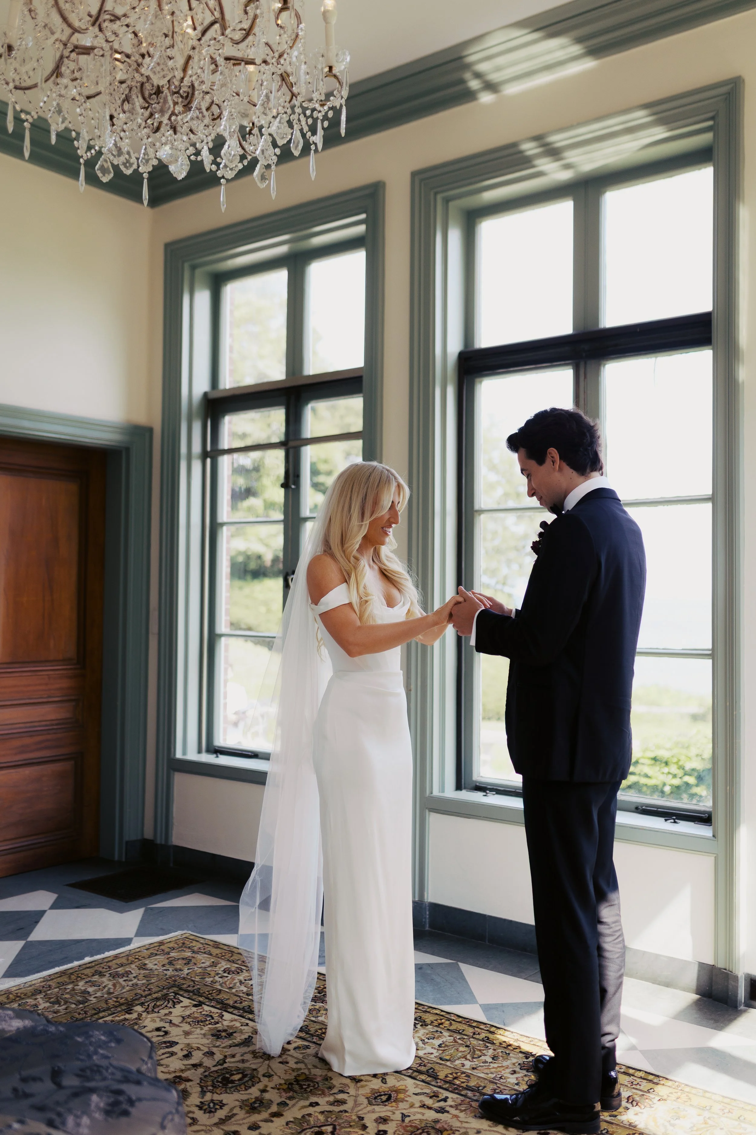 A bride and groom exchange rings inside a room with large green-framed windows, a chandelier, a wooden door, and a patterned rug.