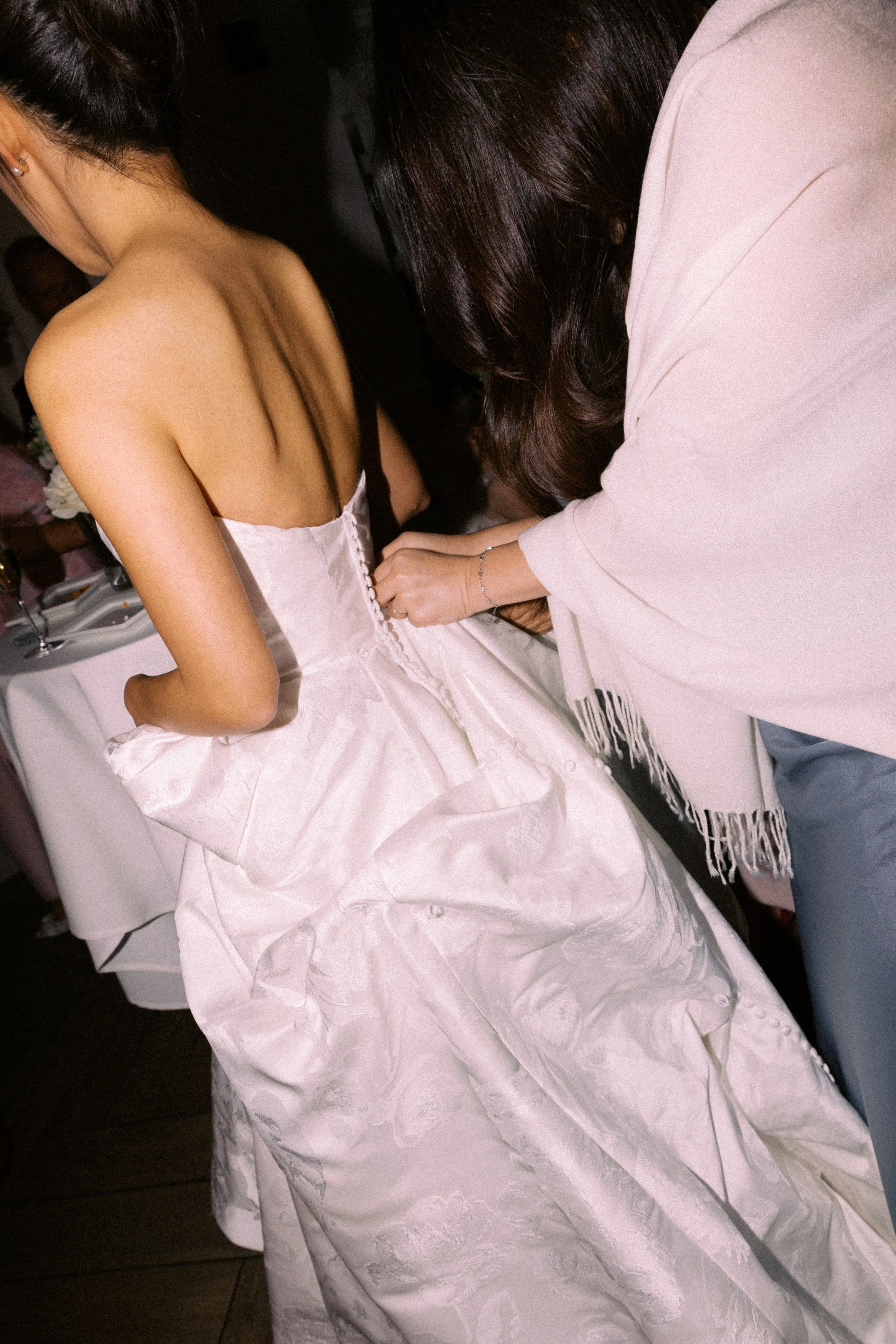 A woman in a strapless white wedding dress being assisted with tying the back buttons by another woman.