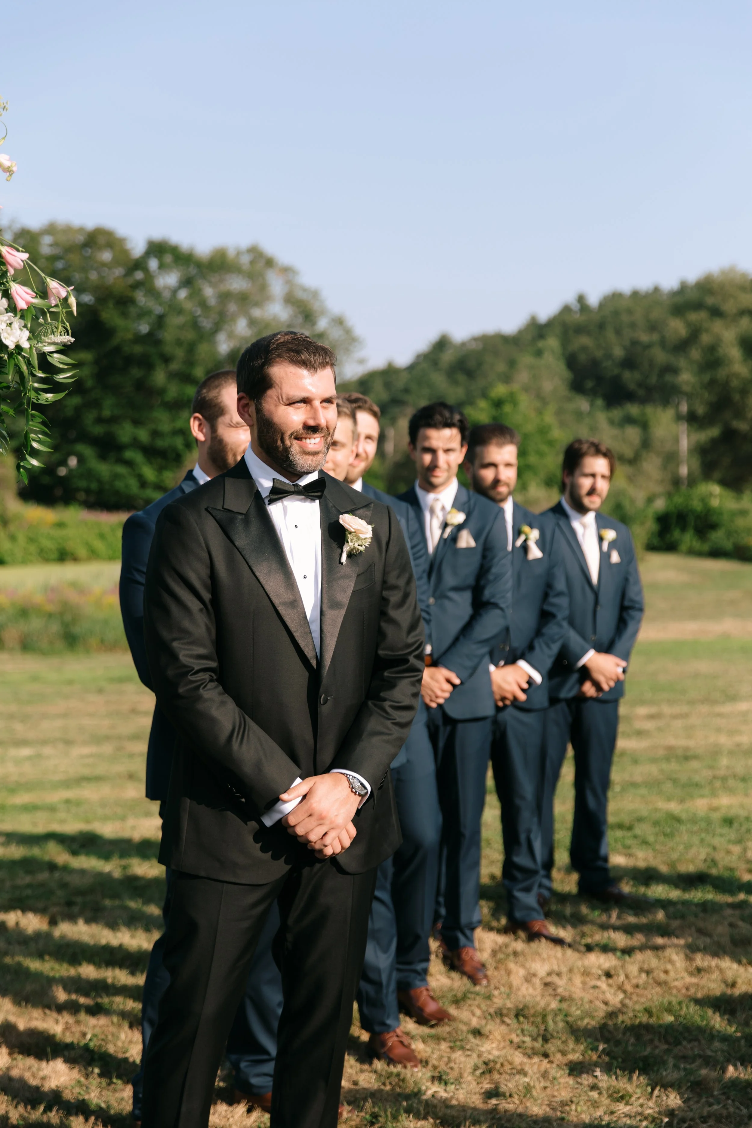 A groom and groomsmen standing outdoors during a wedding ceremony, with the groom in a black tuxedo and the groomsmen in blue suits.
