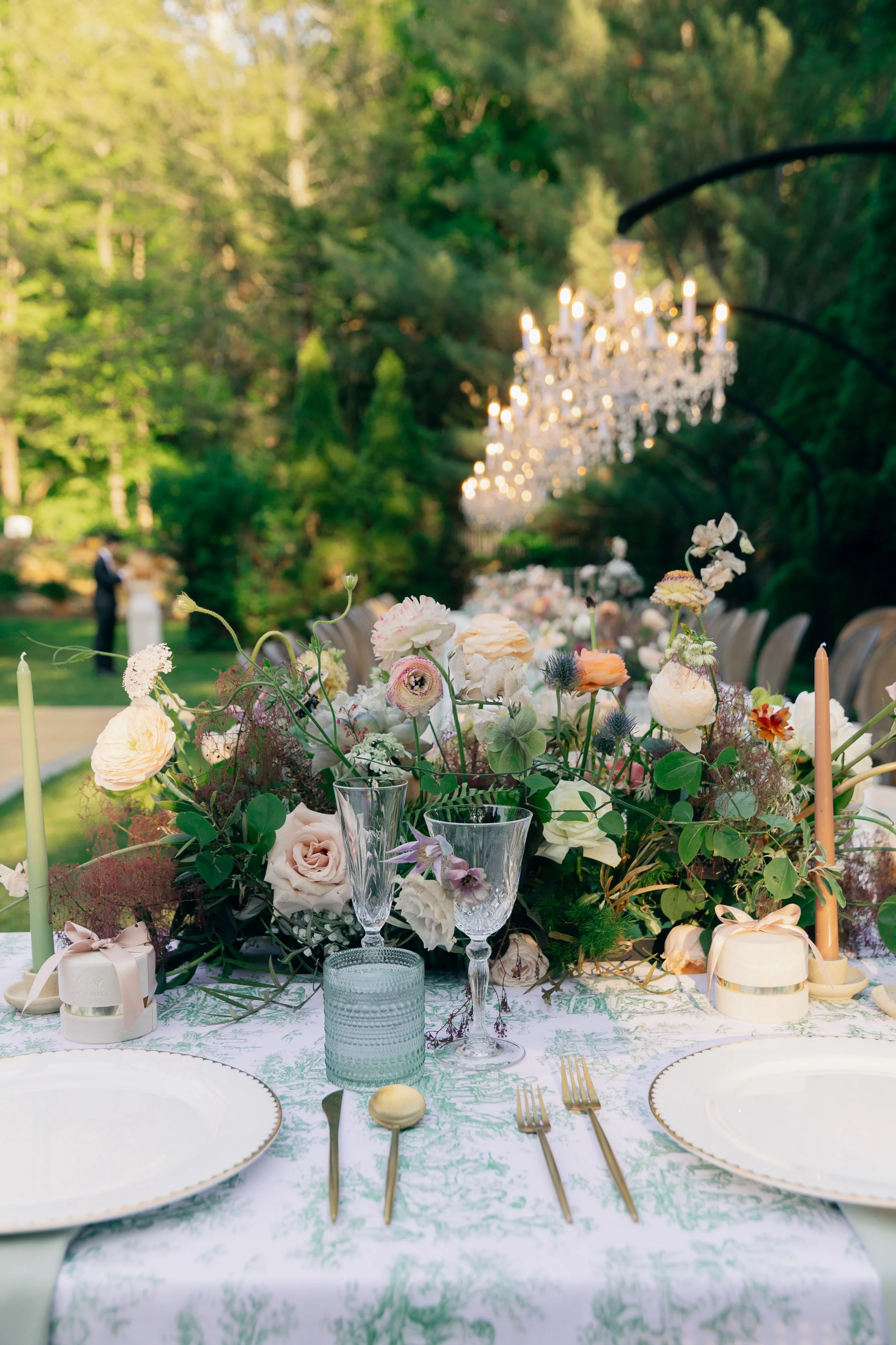 Elegant outdoor wedding table setup with floral centerpiece, glassware, and chandeliers hanging in a lush green garden.