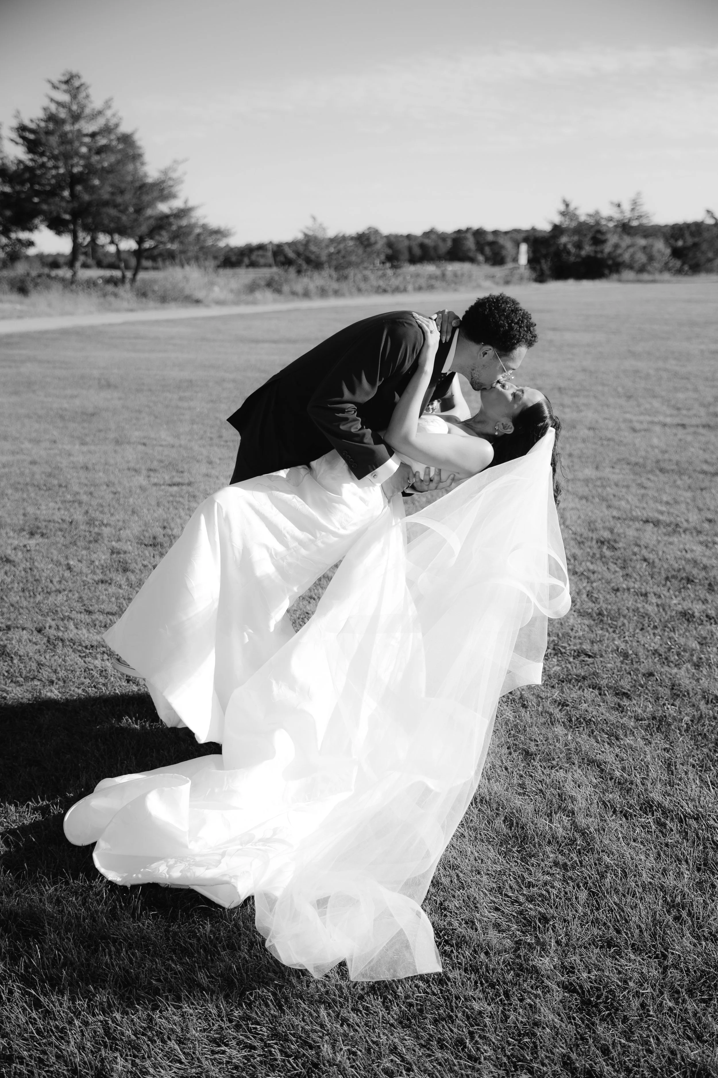 A couple dressed in wedding attire sharing a romantic kiss outdoors on a grassy field.