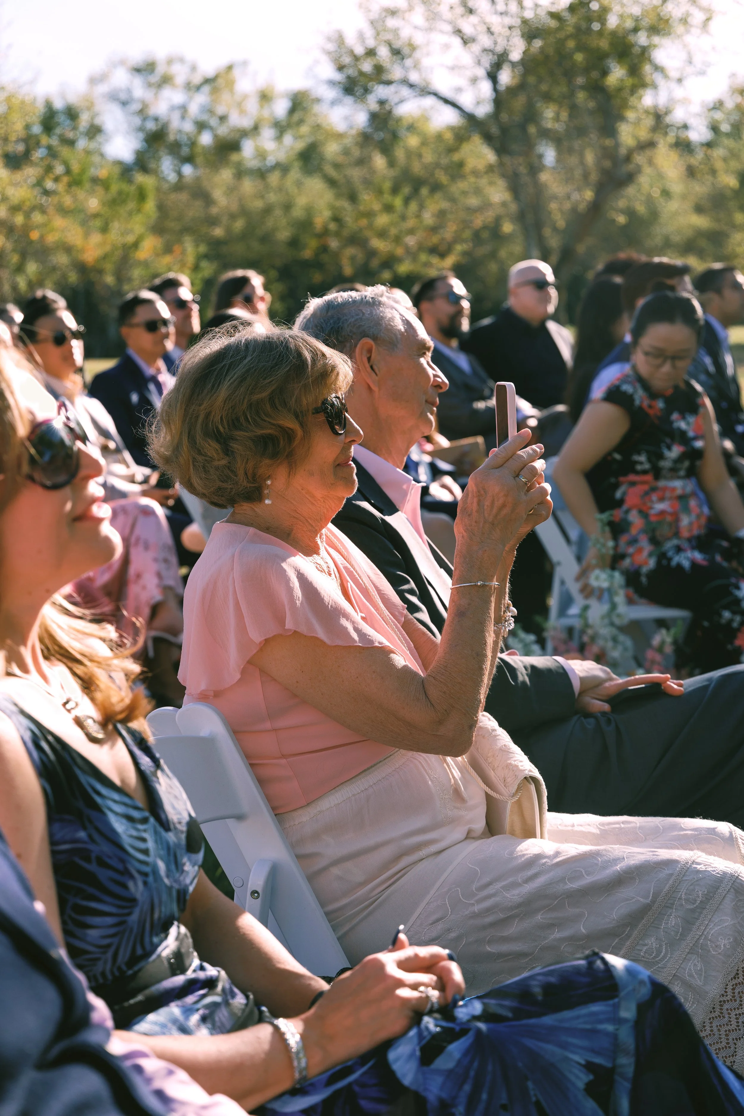 People sitting outdoors attending an event, with some wearing sunglasses and listening attentively, amid trees and sunshine.