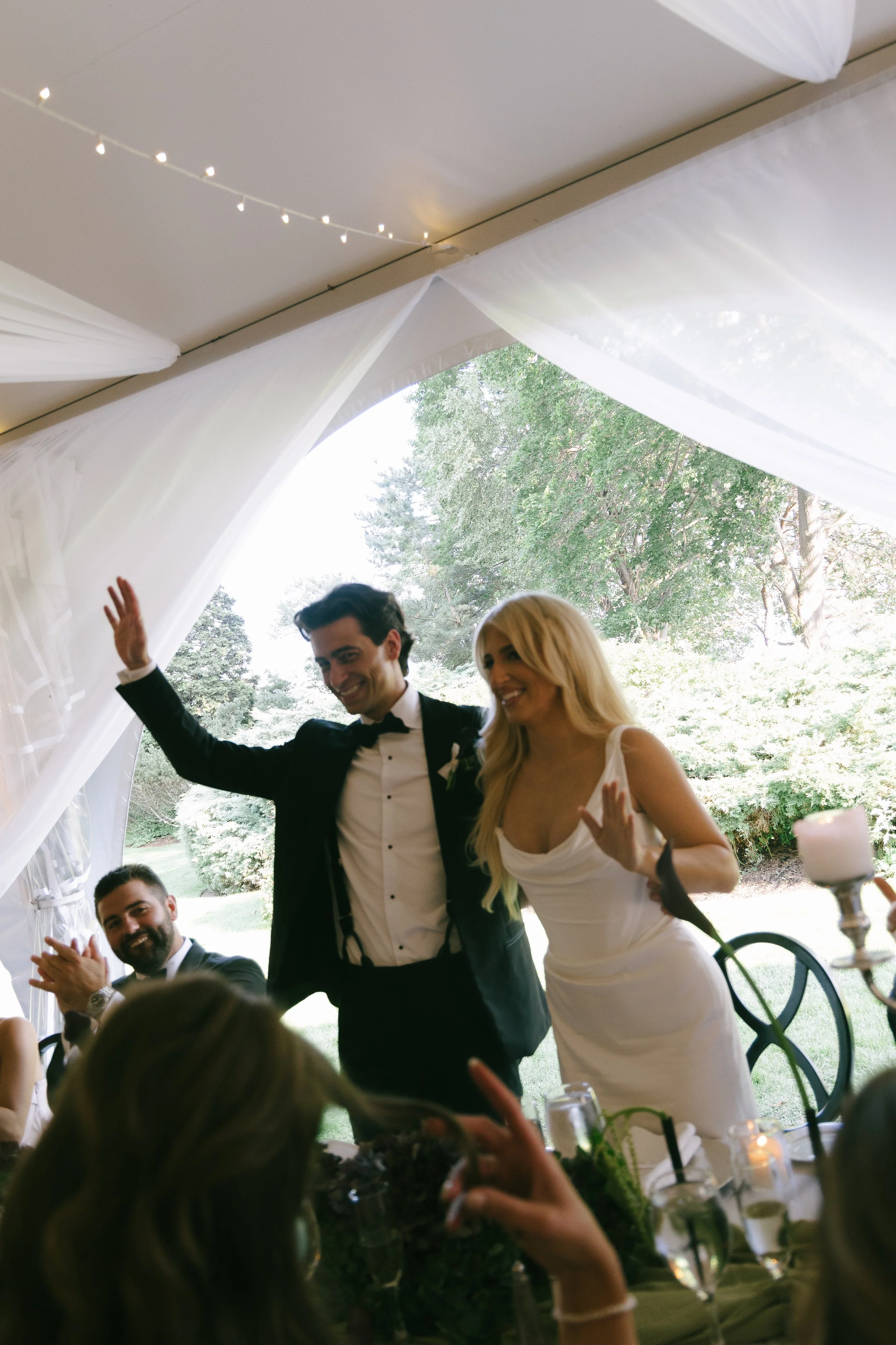 A bride and groom smiling and waving at their wedding reception, with guests seated at a table in front of them. The bride is wearing a white wedding dress and the groom is in a tuxedo. The setting is outdoors with trees visible in the background.