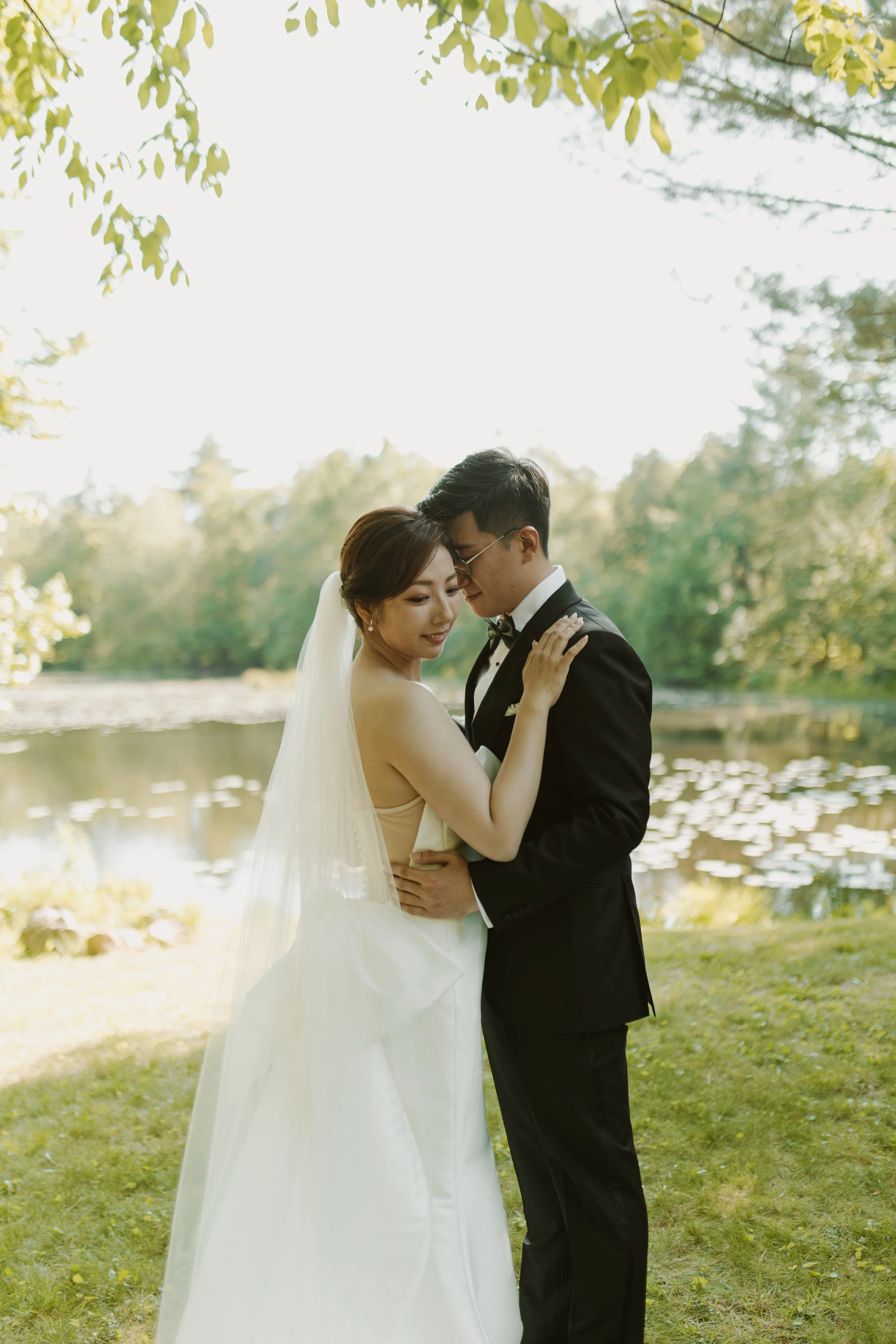 A bride and groom stand close together by a lake at sunset, embracing and looking at each other lovingly.