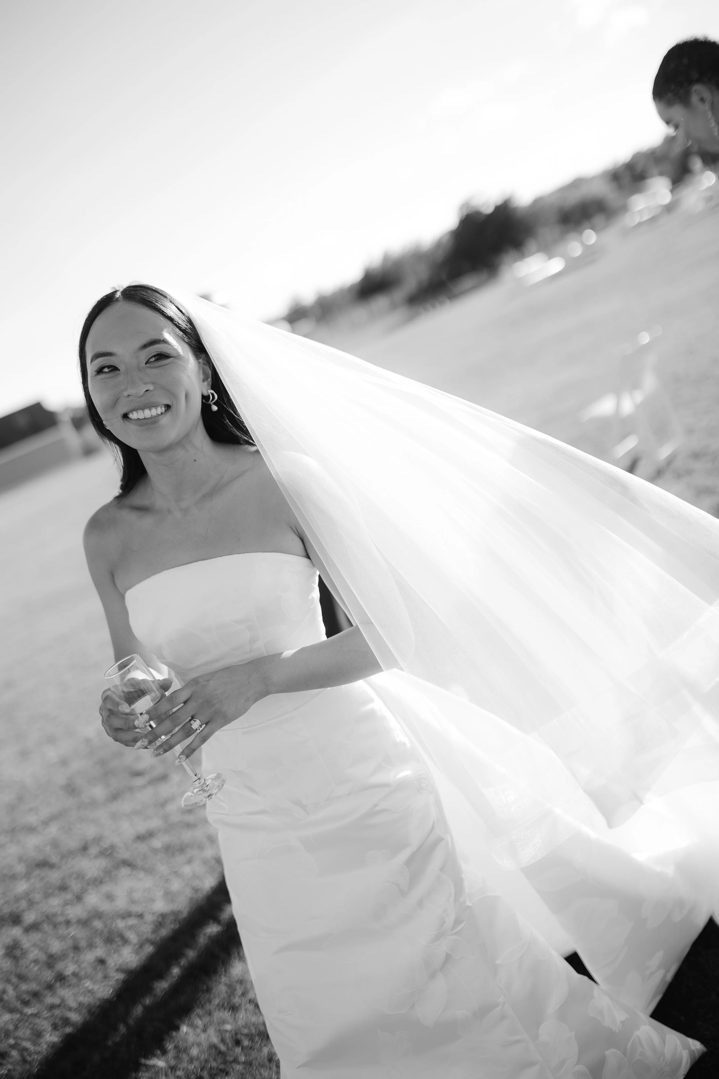 Bride in a strapless wedding dress holding a glass of wine, smiling outdoors in bright sunlight with a flowing veil.
